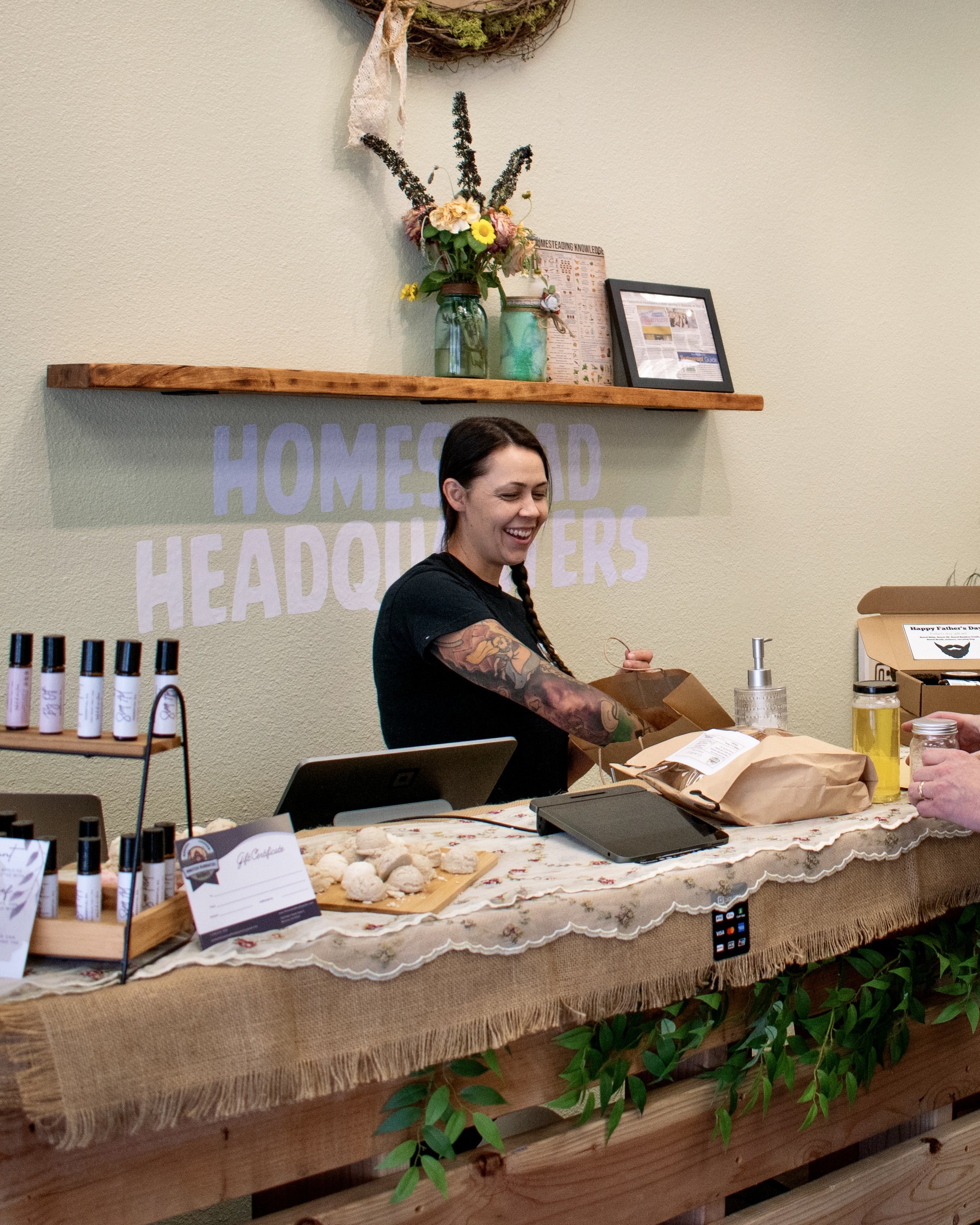 A young woman with a braid and tattoos smiling behind a display table at a market or shop. The table has small bottles, a tablet, and various products on it, with a rustic, decorated backdrop behind her.