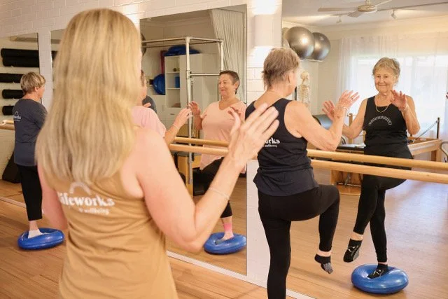 Older women participating in a fitness class using balance boards and a ballet barre in a dance studio.