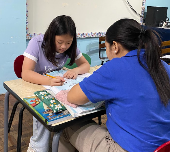 A girl in a purple shirt is doing homework at a desk with a woman in a blue shirt, surrounded by books and school supplies in a classroom.