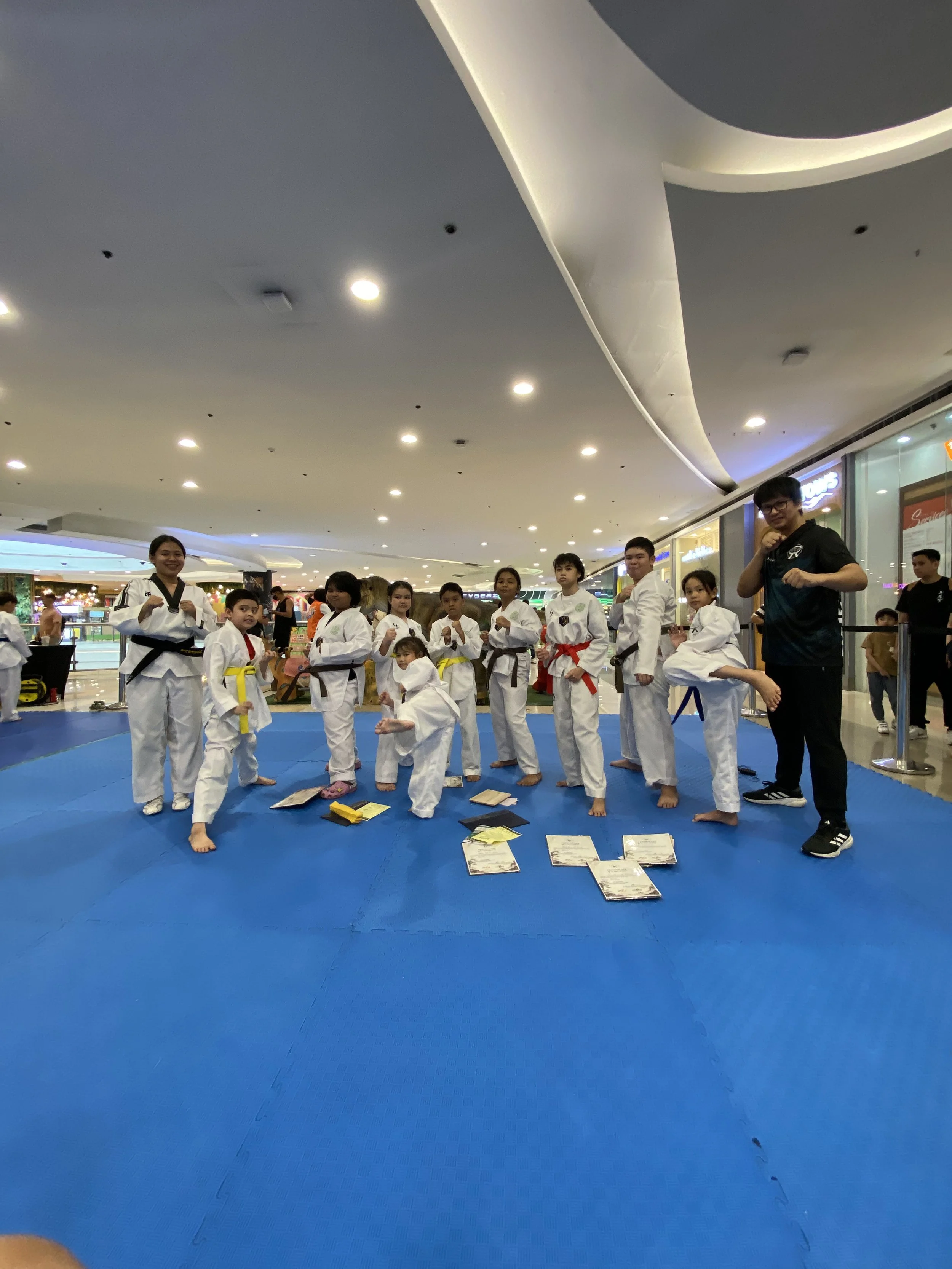 Group of five children and a teenager in martial arts uniforms standing on a mat with arms crossed in a martial arts dojo.