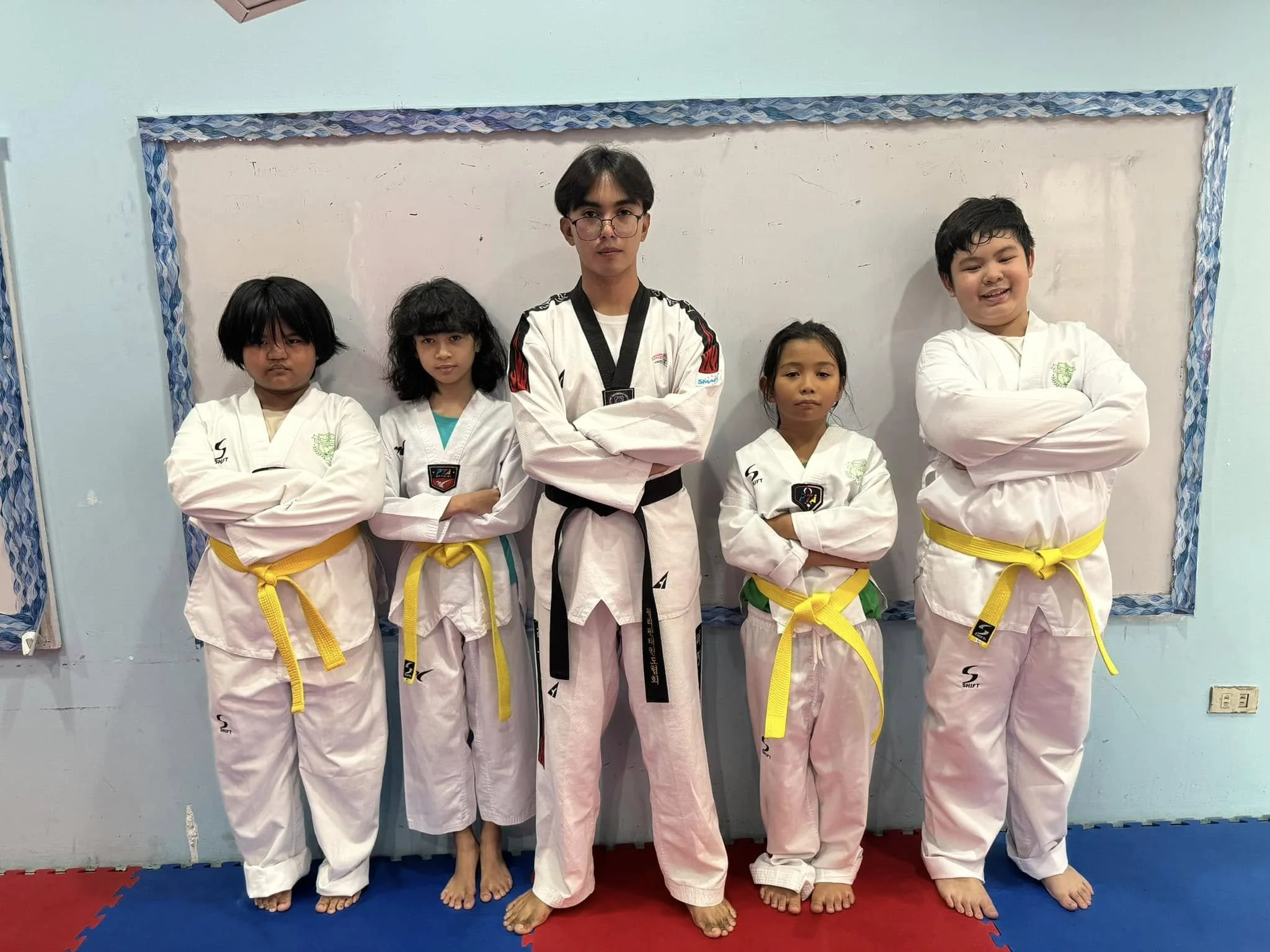 Group of five children and a teenager in martial arts uniforms standing on a mat with arms crossed in a martial arts dojo.