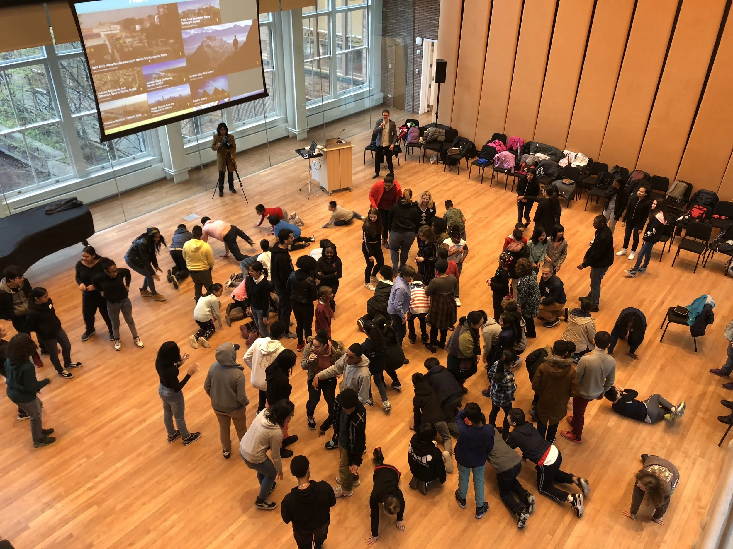 A large group of people, including children and adults, are participating in an indoor activity in a spacious hall with wooden flooring. Some are crawling or lying on the floor, while others are standing and watching. There is a large screen and a woman with a camera on a tripod near the windows, which show a view of trees outside.