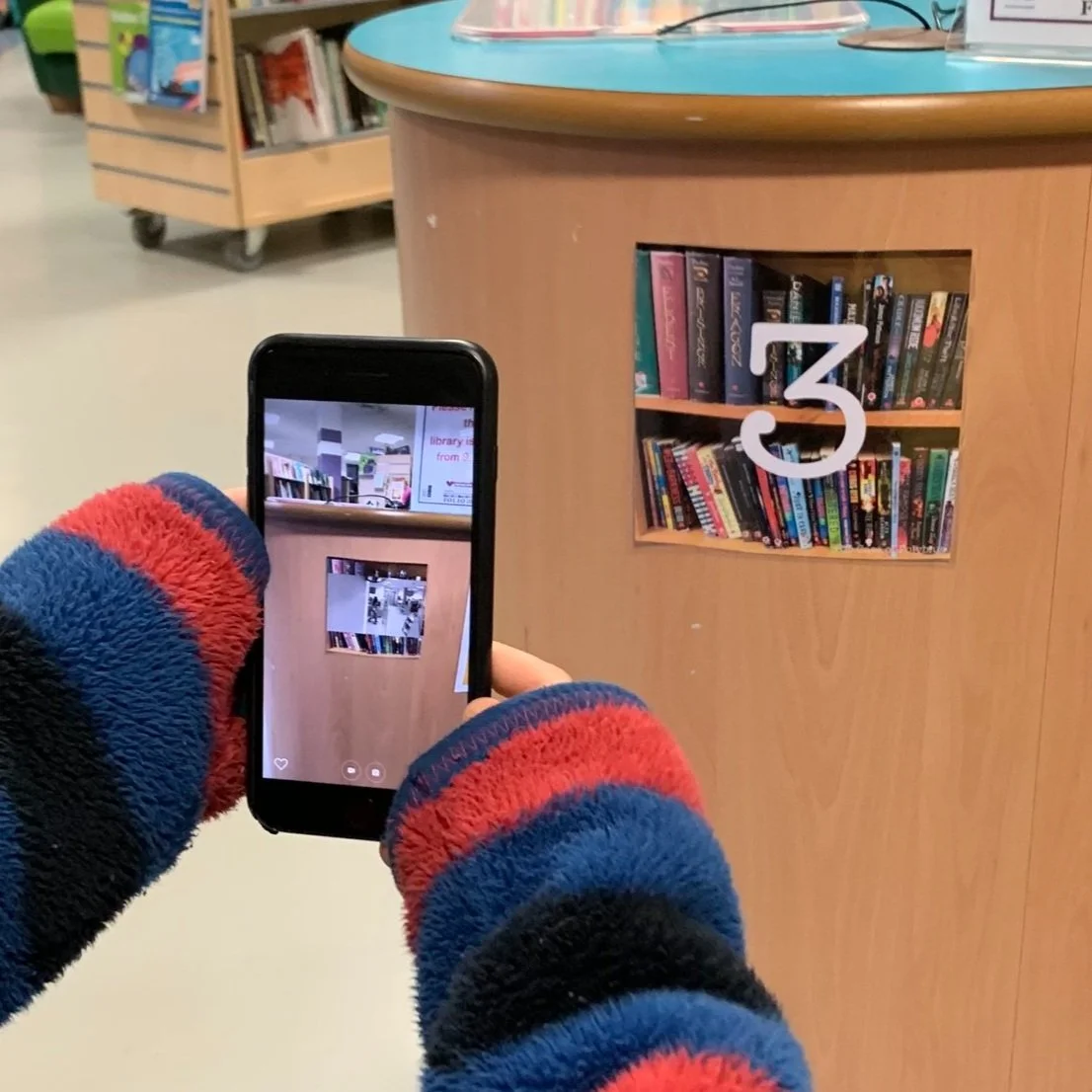 Person taking a photo of a library bookshelf with a phone. The bookshelf is labeled with a large white number 3 and contains various books.
