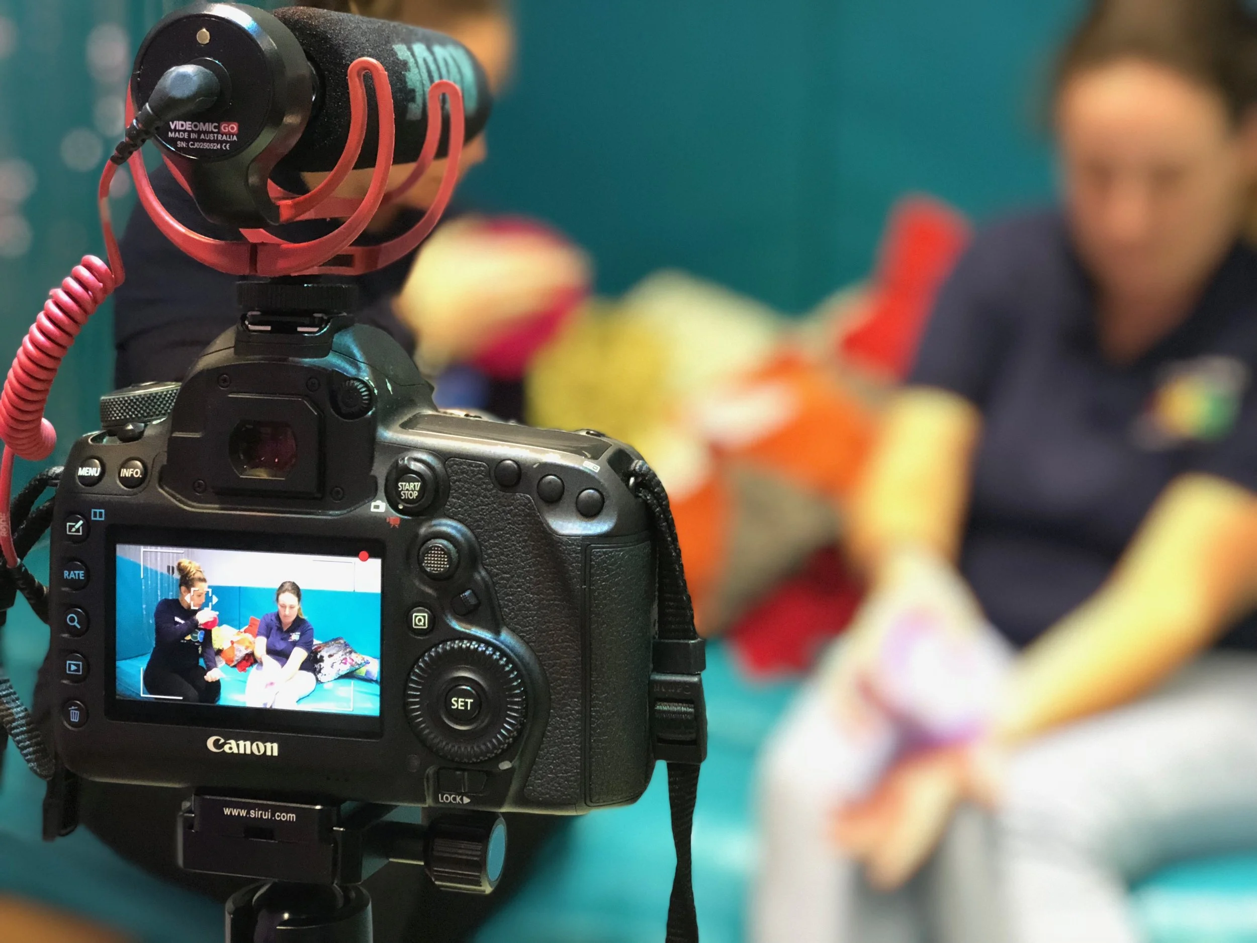 A Canon camera on a tripod capturing a scene of two women sitting on a blue couch, with one woman showing the other a small object. The photo on the camera screen shows this interaction clearly, while the background is blurred.