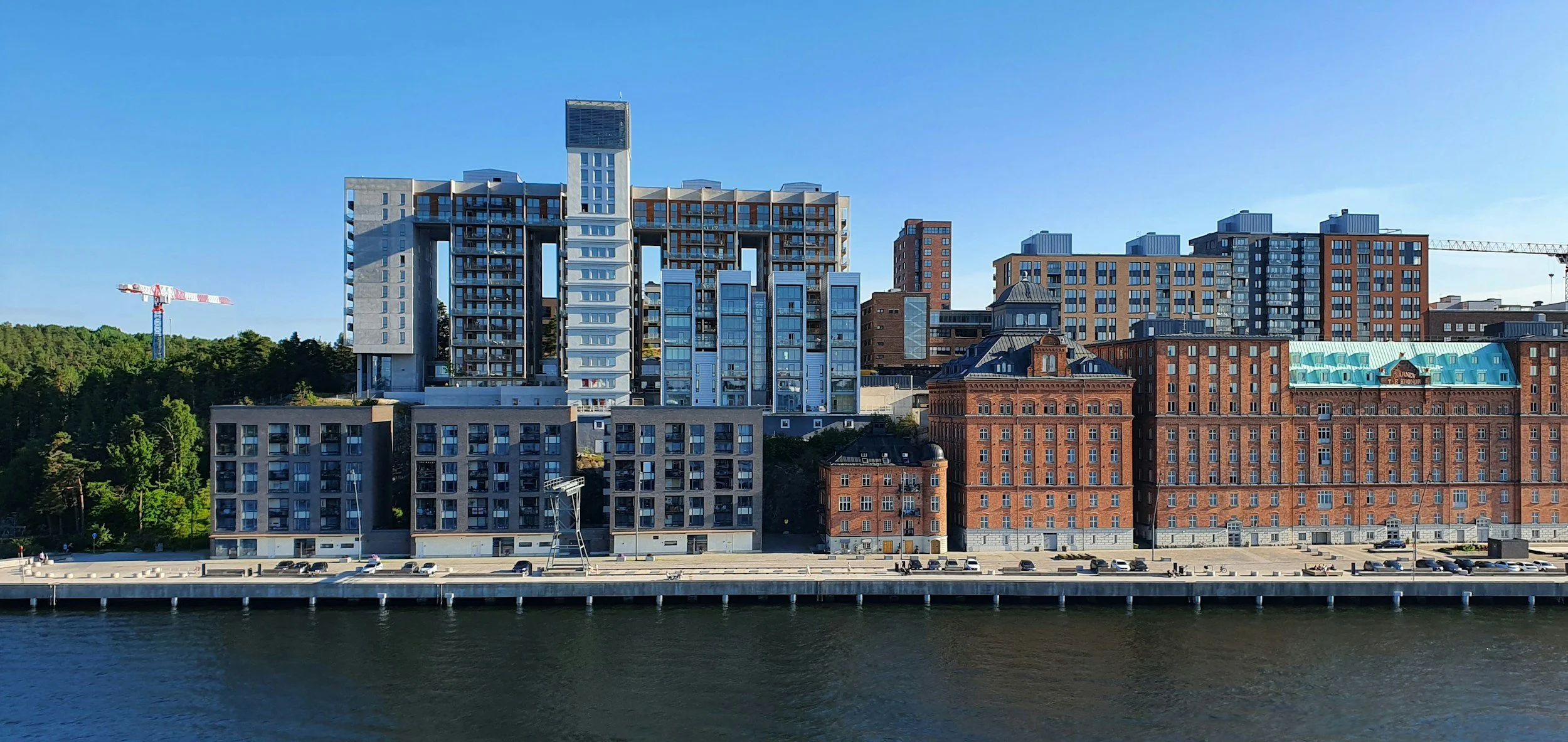 Moderna höghus vid vattnet med grön skog i bakgrunden och en blå klar himmel.