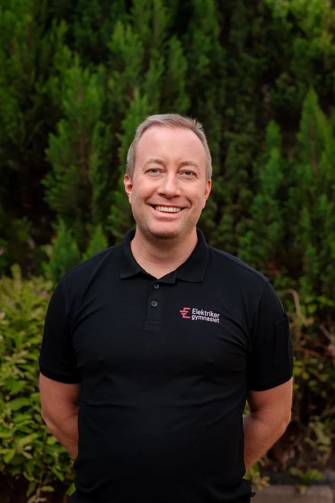 Glad man i svart polo med texten "Elektriker gymnasiet" står utomhus framför gröna buskar.