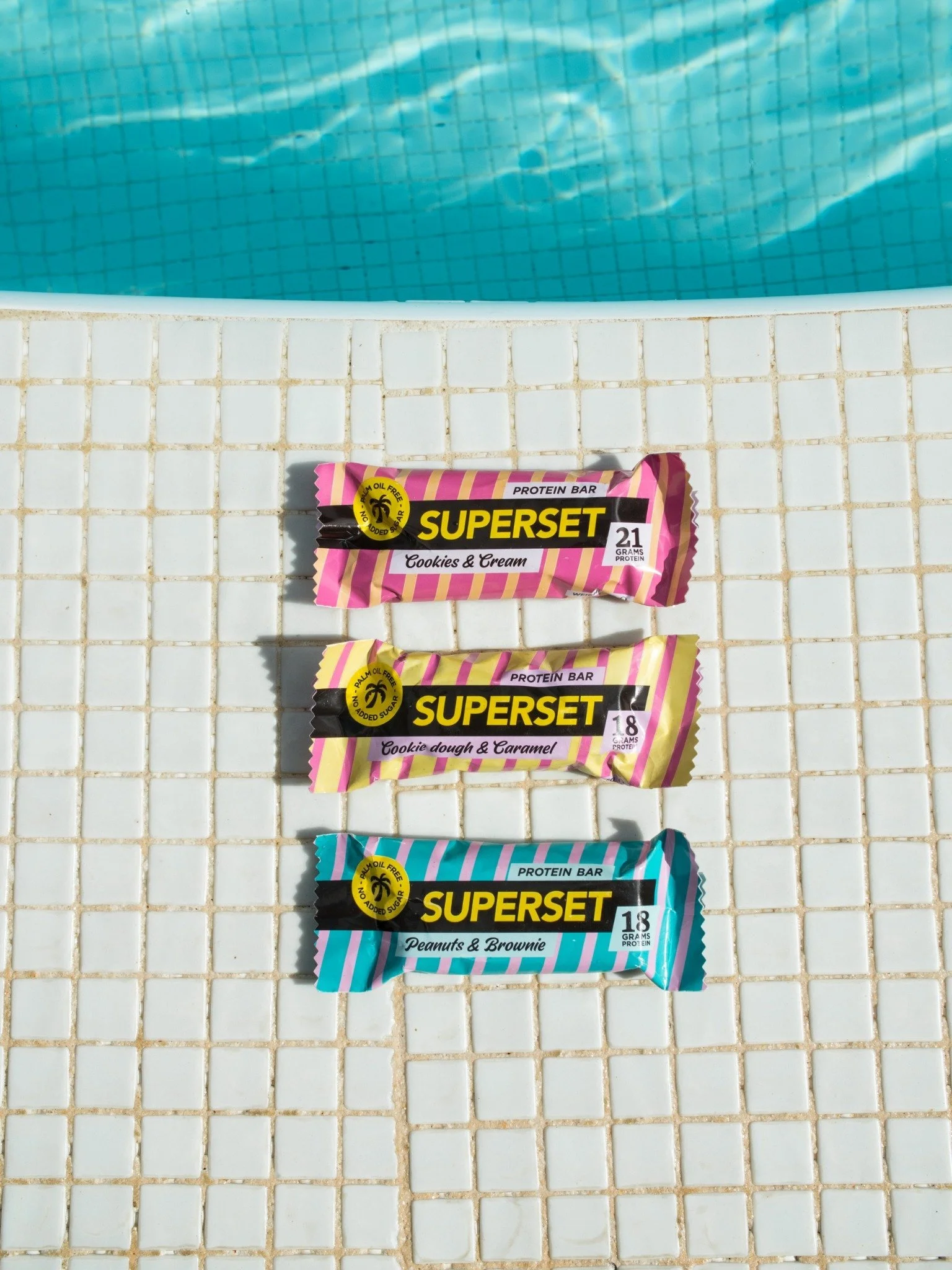 Tre proteinbars i olika färger och smaker placerade på en vit kaklad yta vid poolkanten.