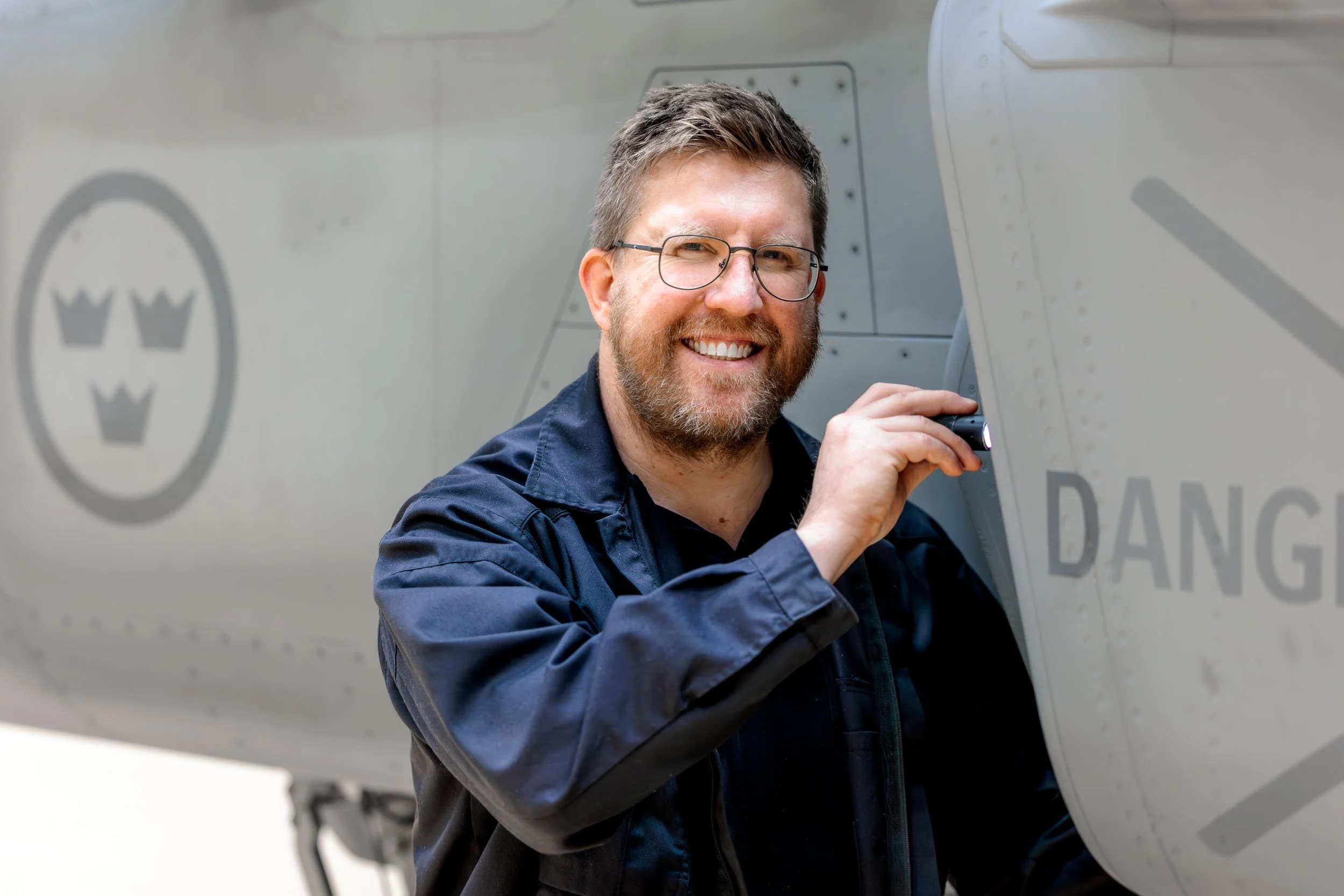 Man med glasögon och skägg som undersöker ett flygplan med ficklampa, ler.