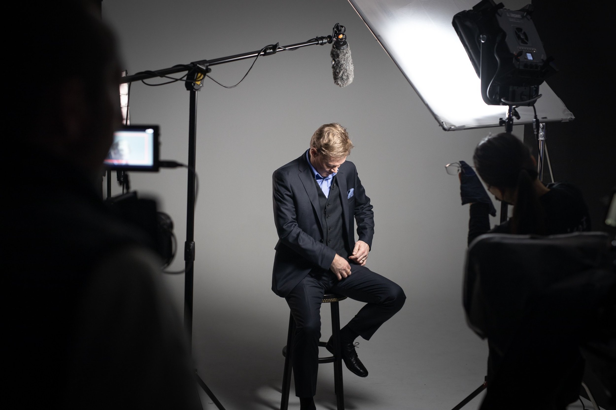 Man i kostym sitter på en stol framför en grå bakgrund under en film- eller fotosession, omgiven av filmcrew med kamera och ljus.