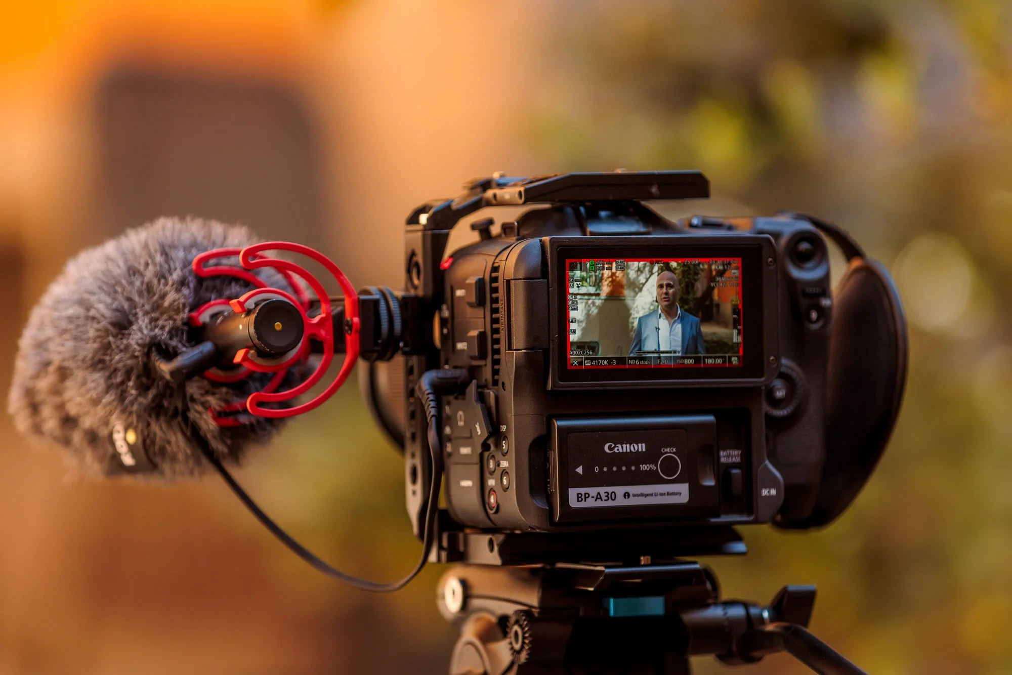 En kamera med en mikrofon på sidan som spelar in ett intervjuporträtt av en man i blå kostym utomhus, med en varm bakgrund av träd och skog.