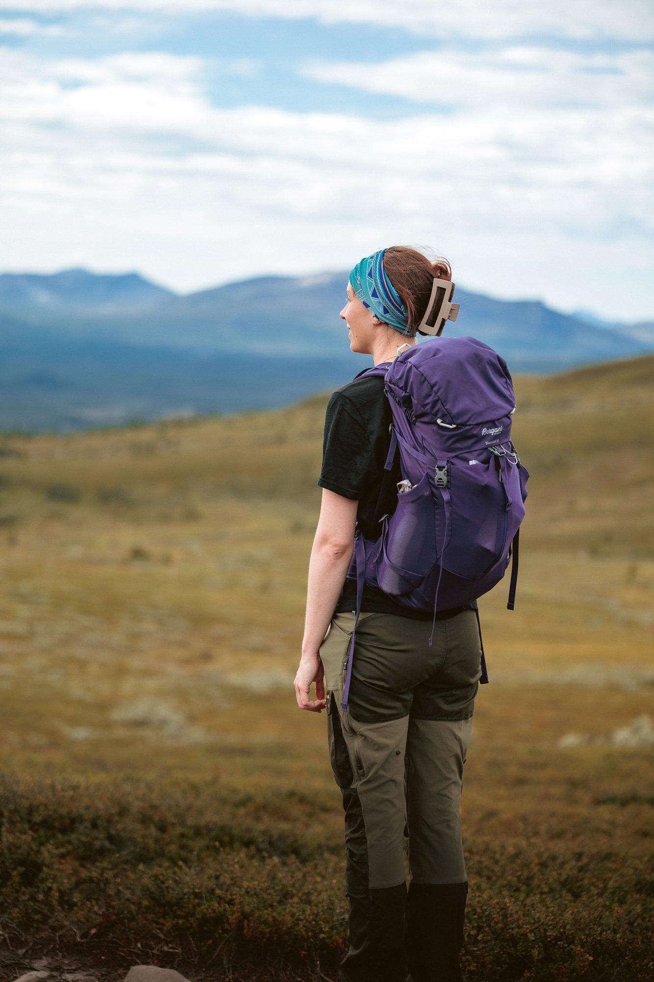 En kvinna med ryggsäck som tittar ut över ett öppet landskap med berg i bakgrunden.
