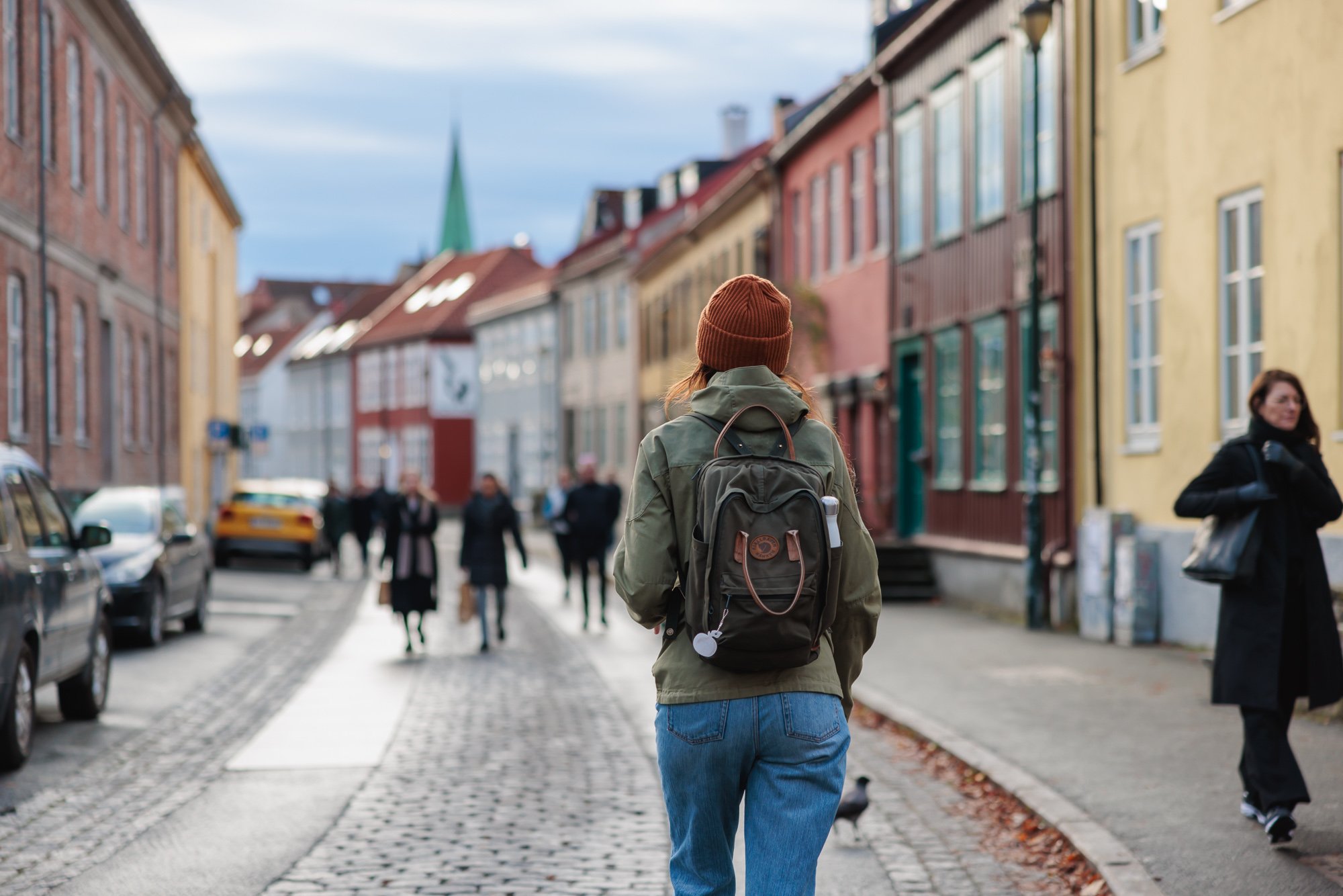 En person i grön jacka och orange mössa med ryggsäck som går längs en gatsträckning med färgglada hus på sidorna, andra människor promenerar.