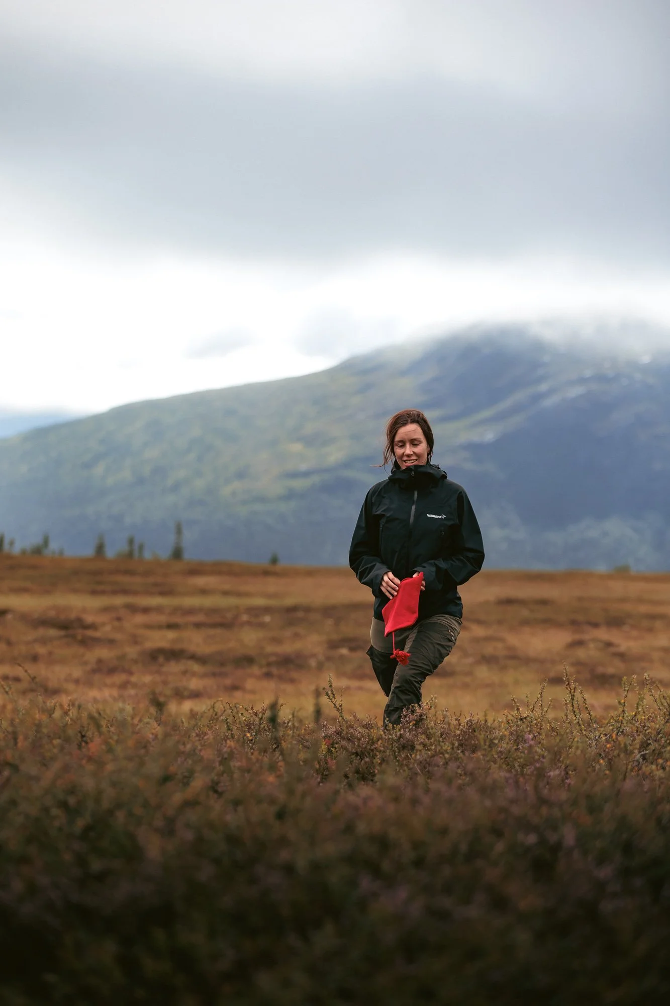 Kvinna går i ett öppet landskap med höga berg i bakgrunden, bär en svart jacka och håller i en liten röd påse.