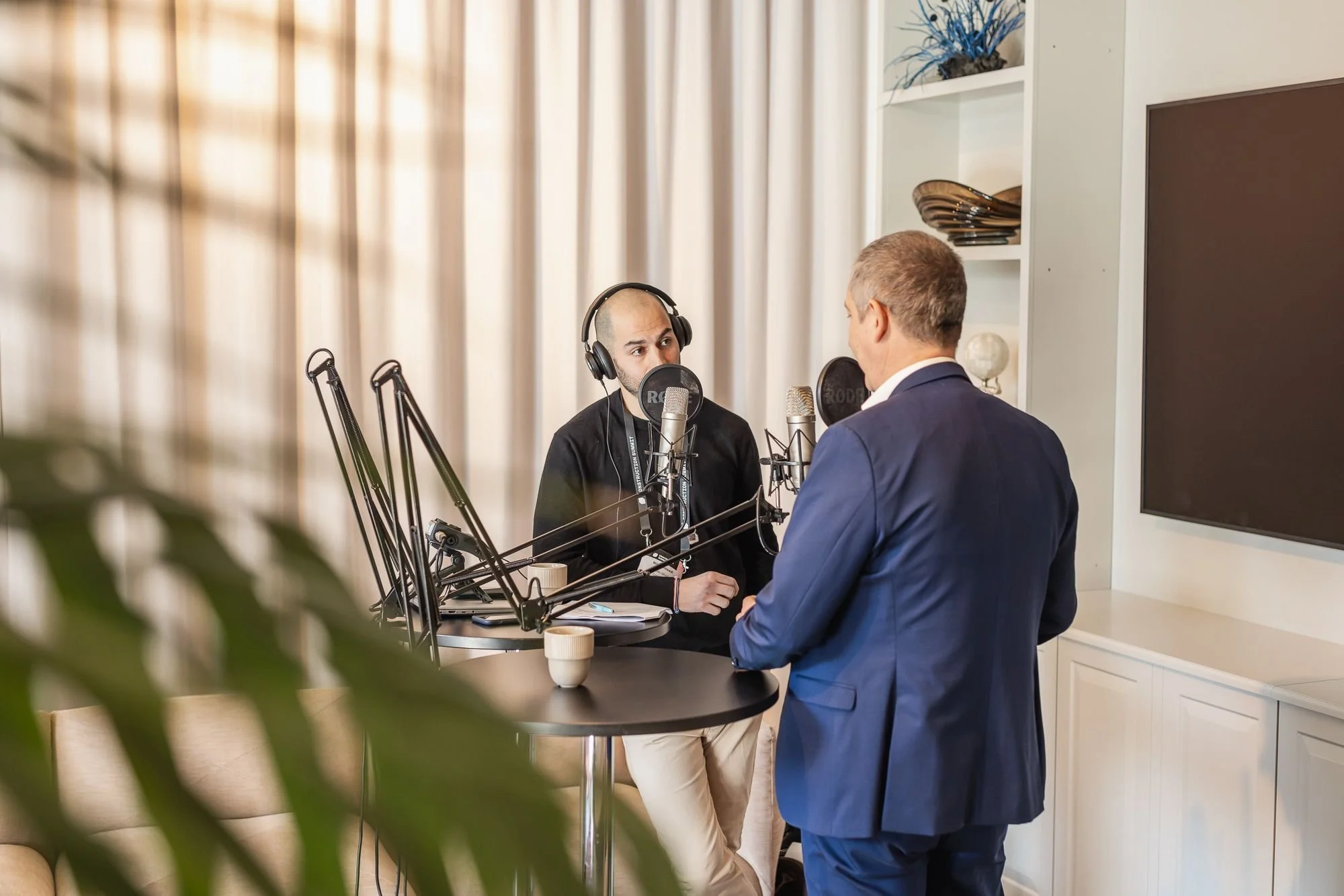 Två män engagerade i en inspelning i en studio, en med hörlurar framför mikrofon och en annan i kostym, samtal.