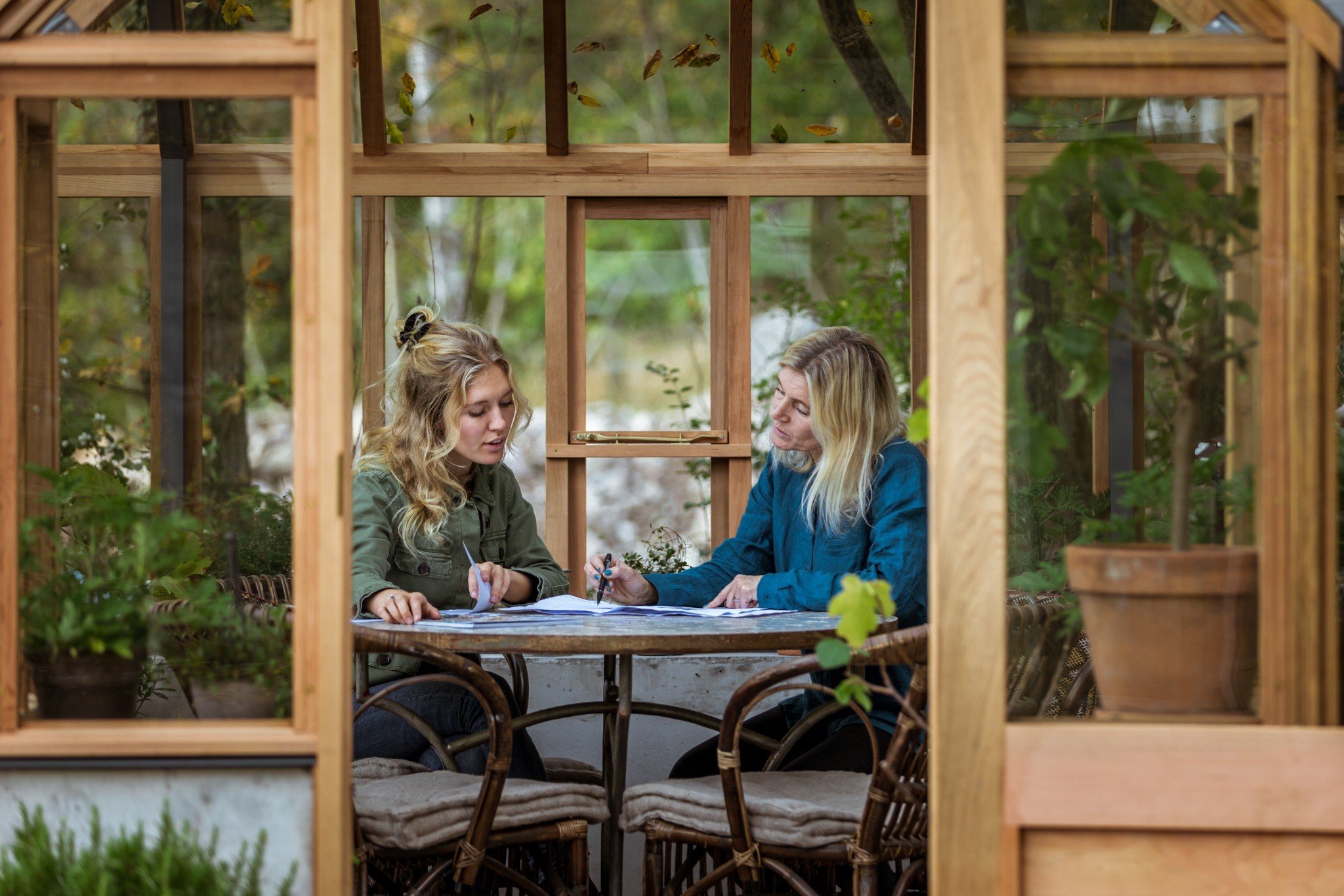 Två kvinnor sitter vid ett bord och diskuterar utomhus, omgiven av en träpergola och grönt lövverk.