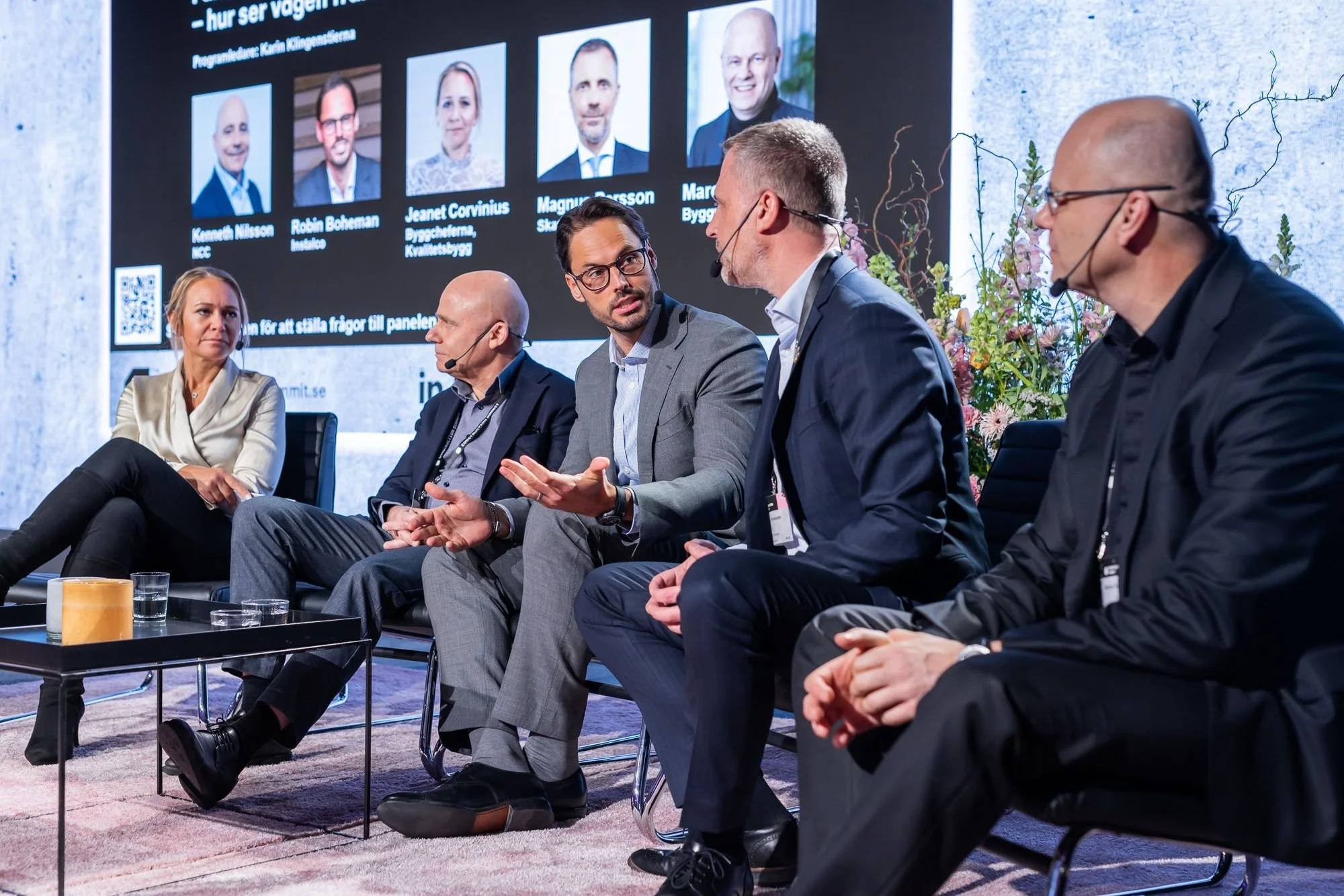 Fem personer deltar i en paneldiskussion på en konferens. En man är i mitten och pratar, medan de andra lyssnar. En stor skärm i bakgrunden visar bilder av paneldeltagarna och text på svenska.