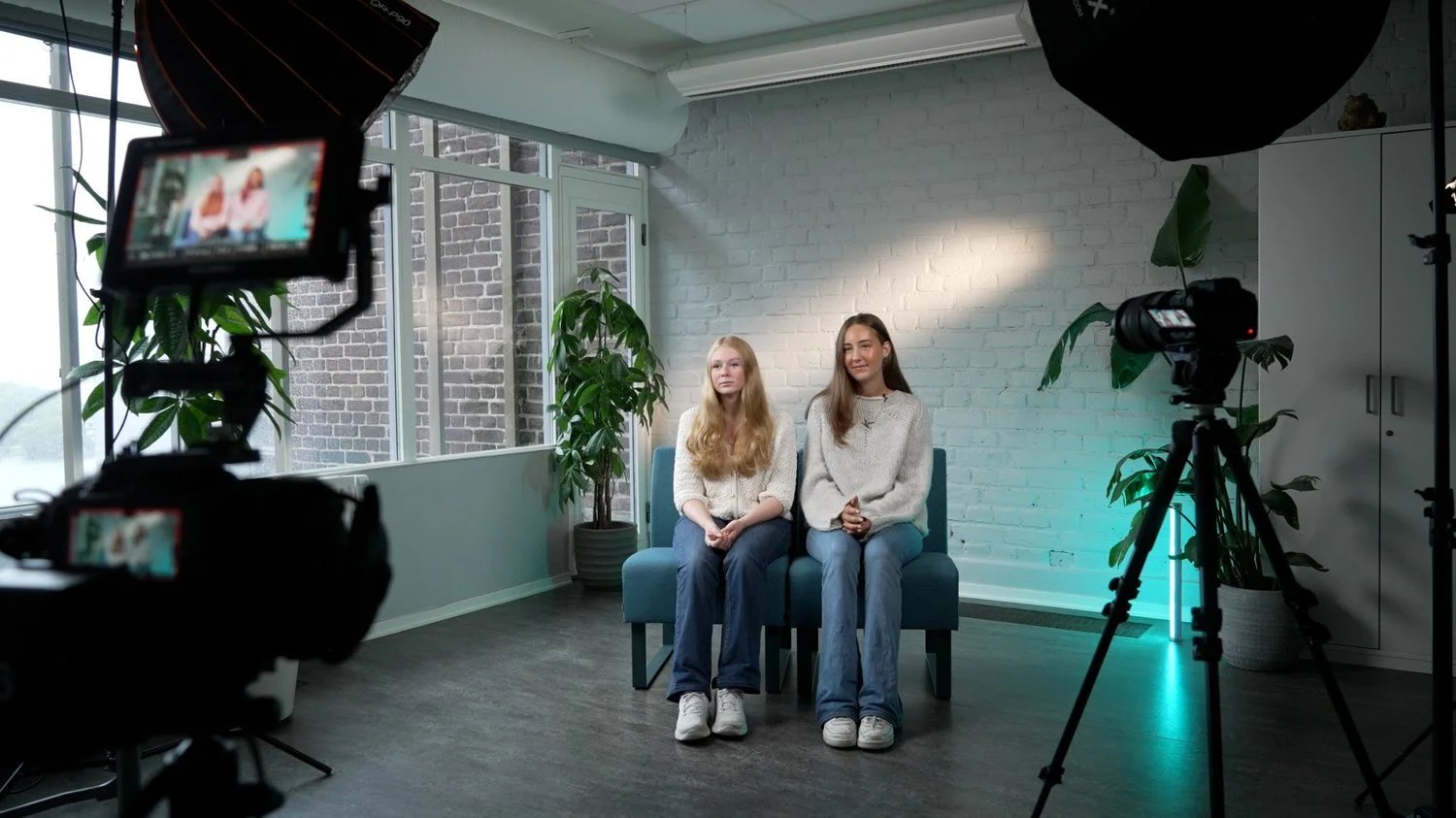 Två unga kvinnor sitter på en blå soffa i ett ljust rum med vita väggar, bakom dem finns ett fönster och gröna växter. De är omgivna av videoutrustning för ett videoinspelning.