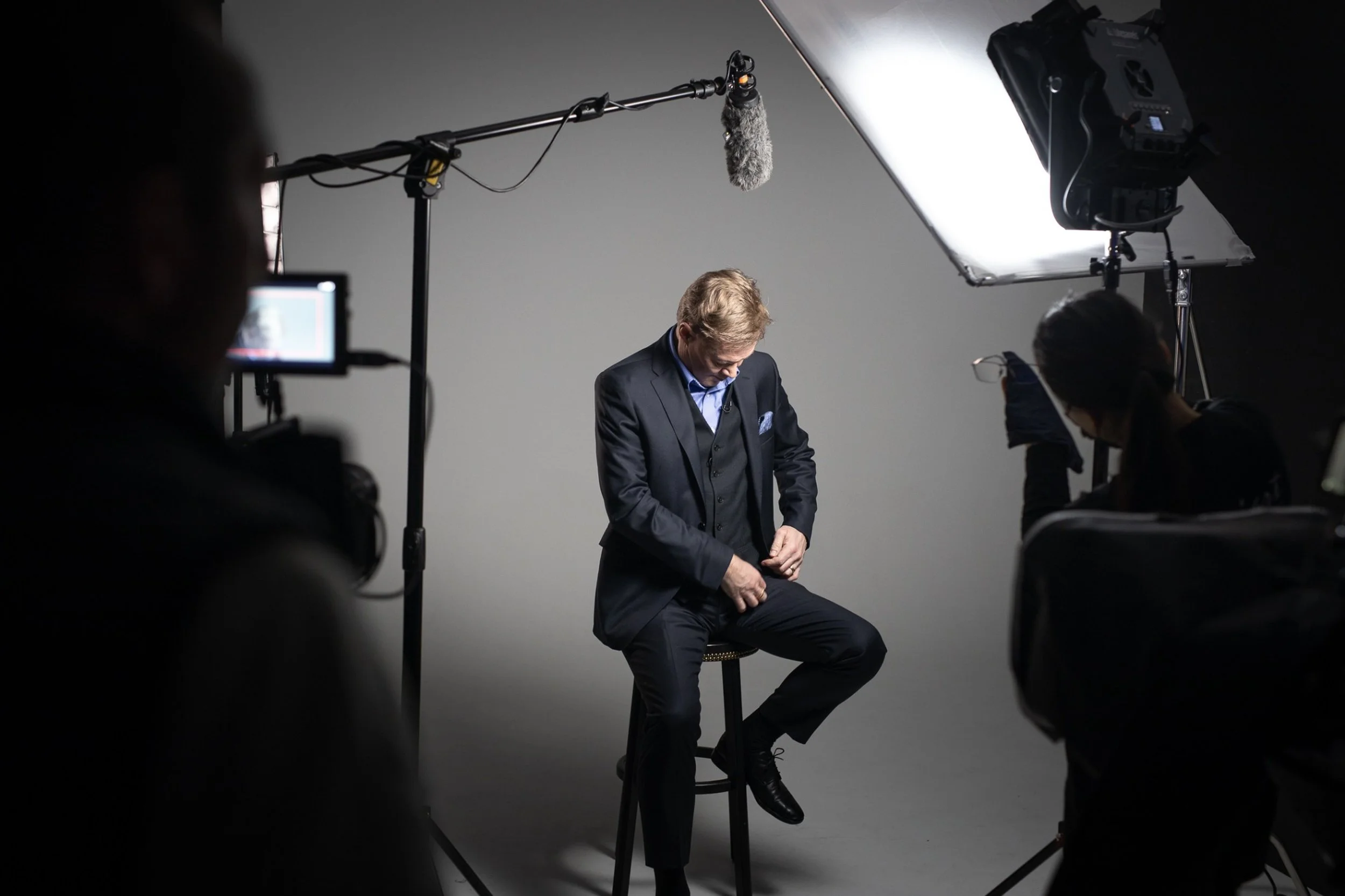Man i kostym sitter på en stol inför en filminspelning eller fotografering, omgivet av fotograf- och filmskapare, i en studio med grå bakgrund.