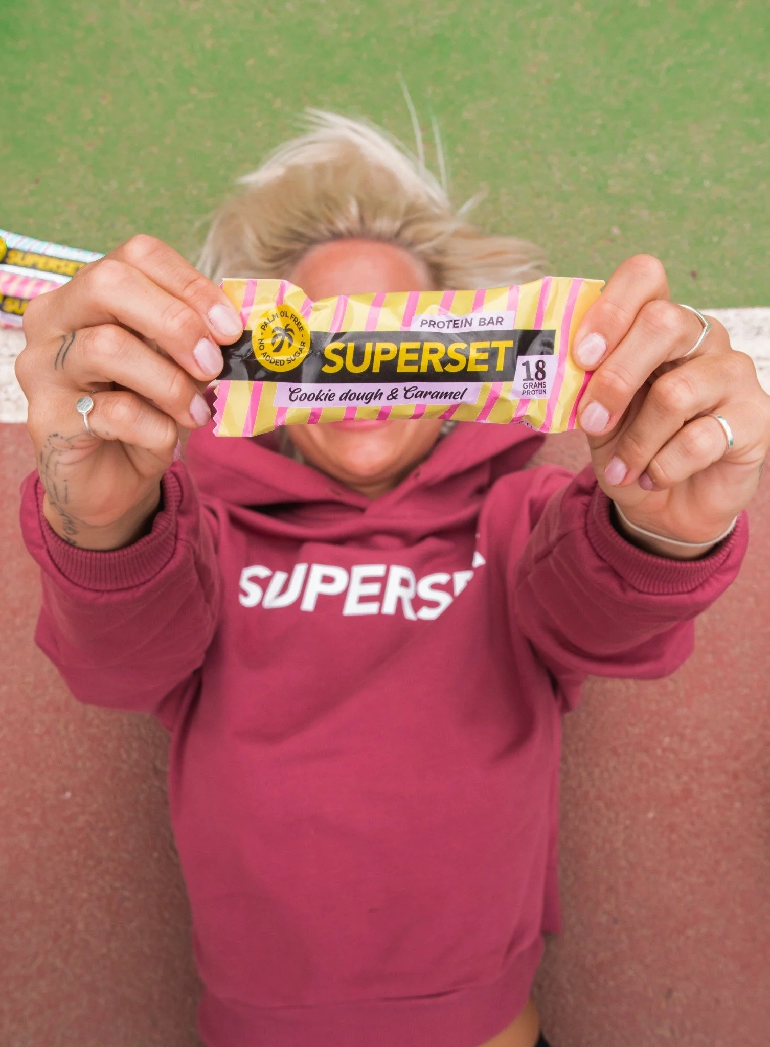En person i en rosa hoodie med texten 'SUPERS' håller en proteinbar framför ansiktet. Bakgrunden är en grön och rosa yta.
