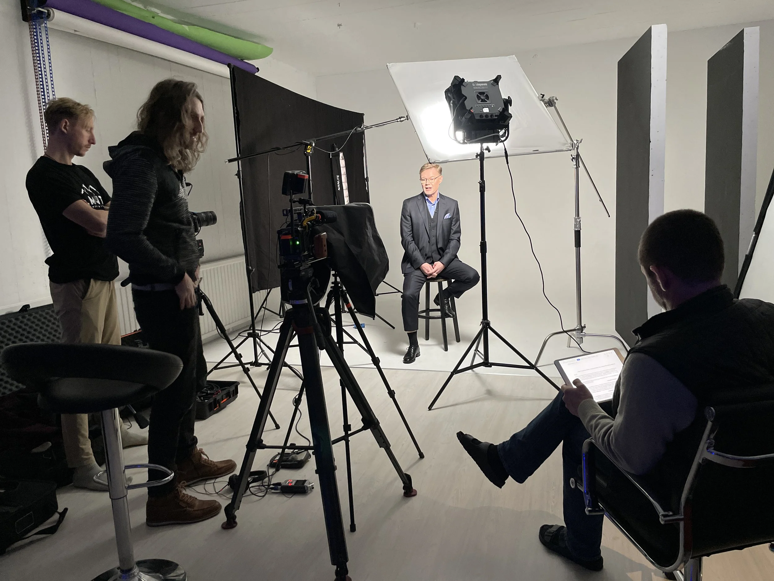 Person som sitter på en stol i ett professionellt fotostudio medan filmteam förbereder inspelning med utrustning som kameror, lampor och ljudutrustning. Personer står och tittar på, en av dem använder en surfplatta.