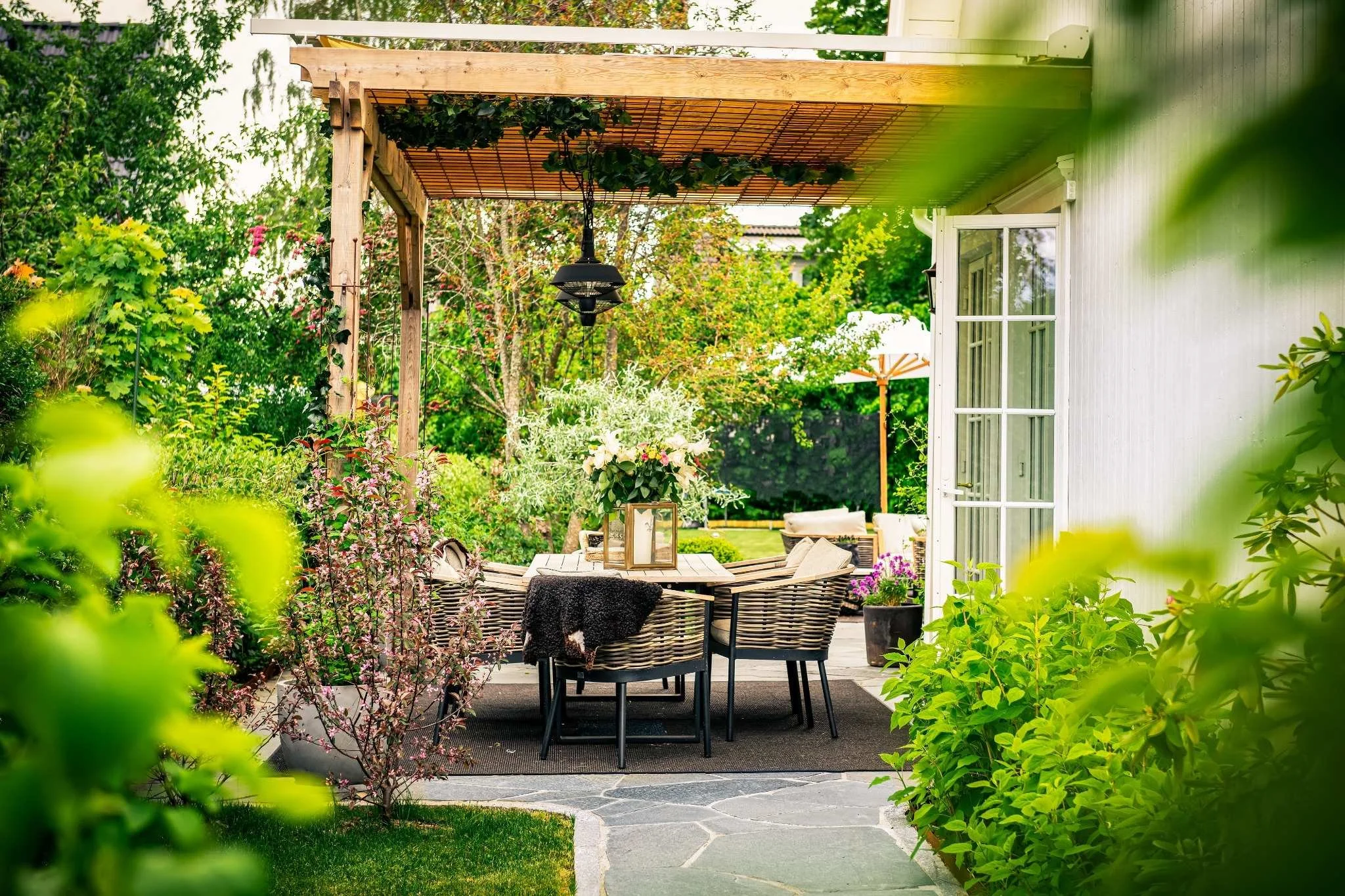 En vacker uteplats med ett runt bord och sex stolar, omgiven av gröna buskar och träd, samt en pergola med tak av trä. Det finns blomsterarrangemang på bordet och en parasoll i bakgrunden.