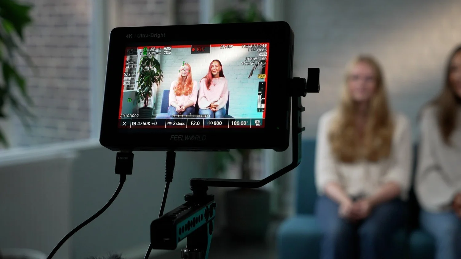 Två kvinnor som sitter på en soffa under en videoinspelning. Skärmen visar att de spelas in i ett studio- eller kontorsmiljö med gröna växter och en vit vägg.