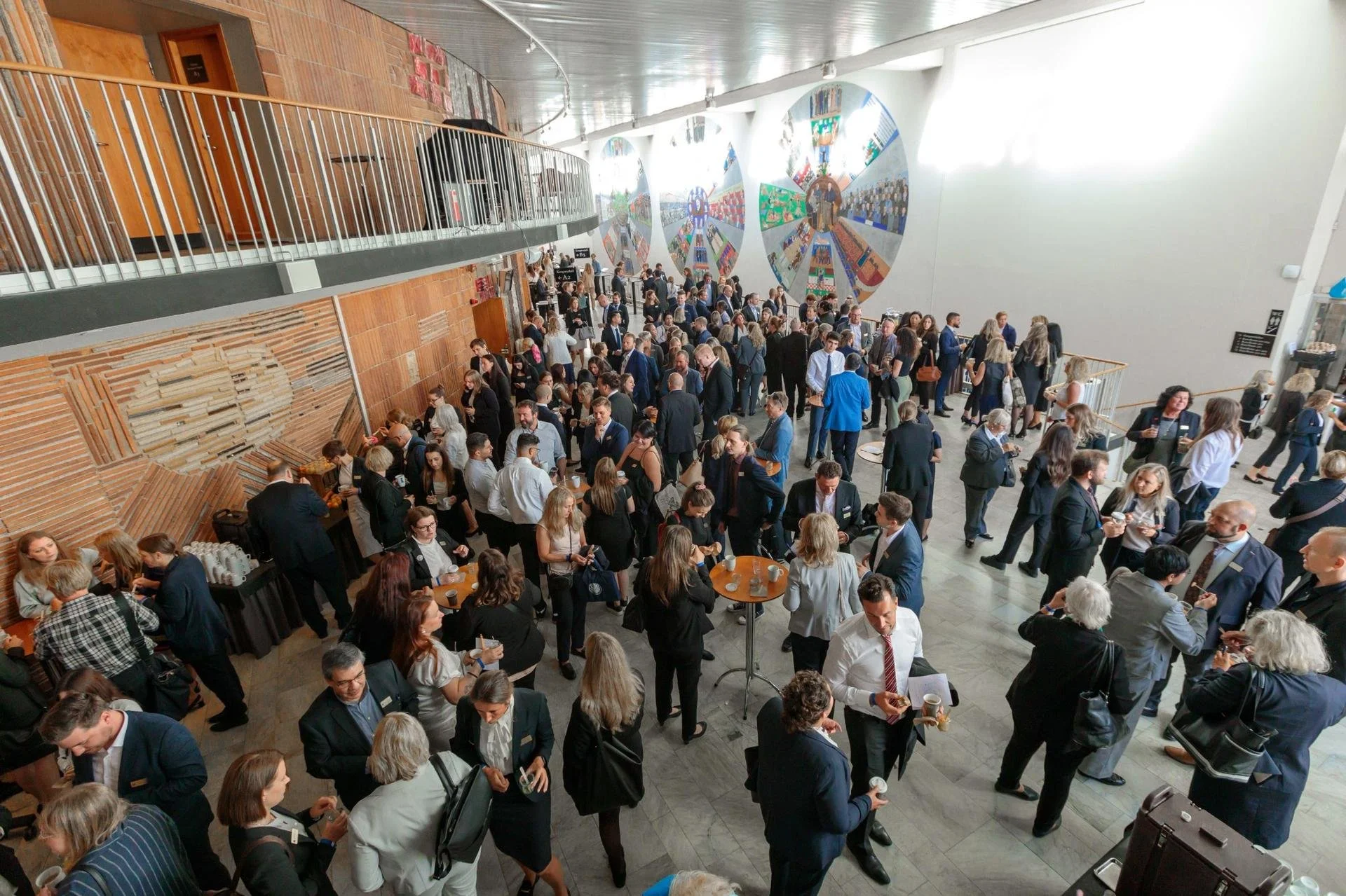 En stor grupp personer vid ett evenemang i en modern inomhuslokal, vissa står i kö för kaffe och minglar, många pratar och leker nätverk. Väggarna är dekorerade med stora, färgglada konstverk.