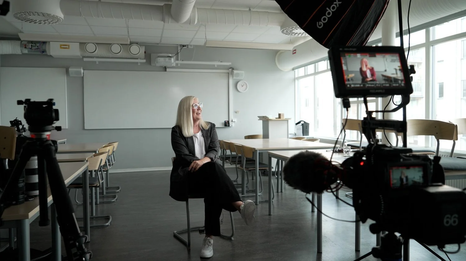 En kvinna med blont hår och glasögon som sitter i ett tomt konferensrum och pratar, inspelad med en videokamera.