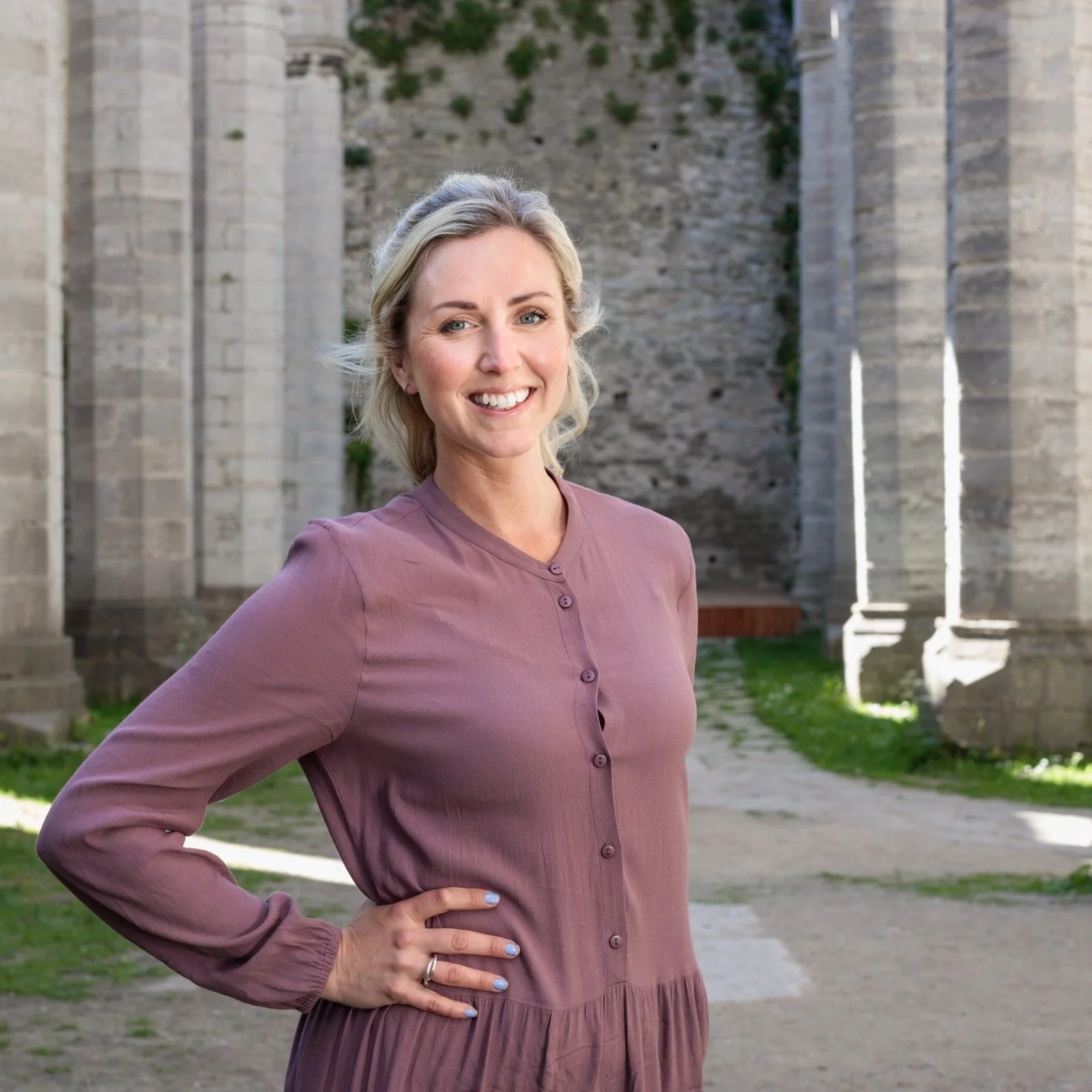 En leende kvinna står utomhus framför ruiner av gamla stenbyggnader.