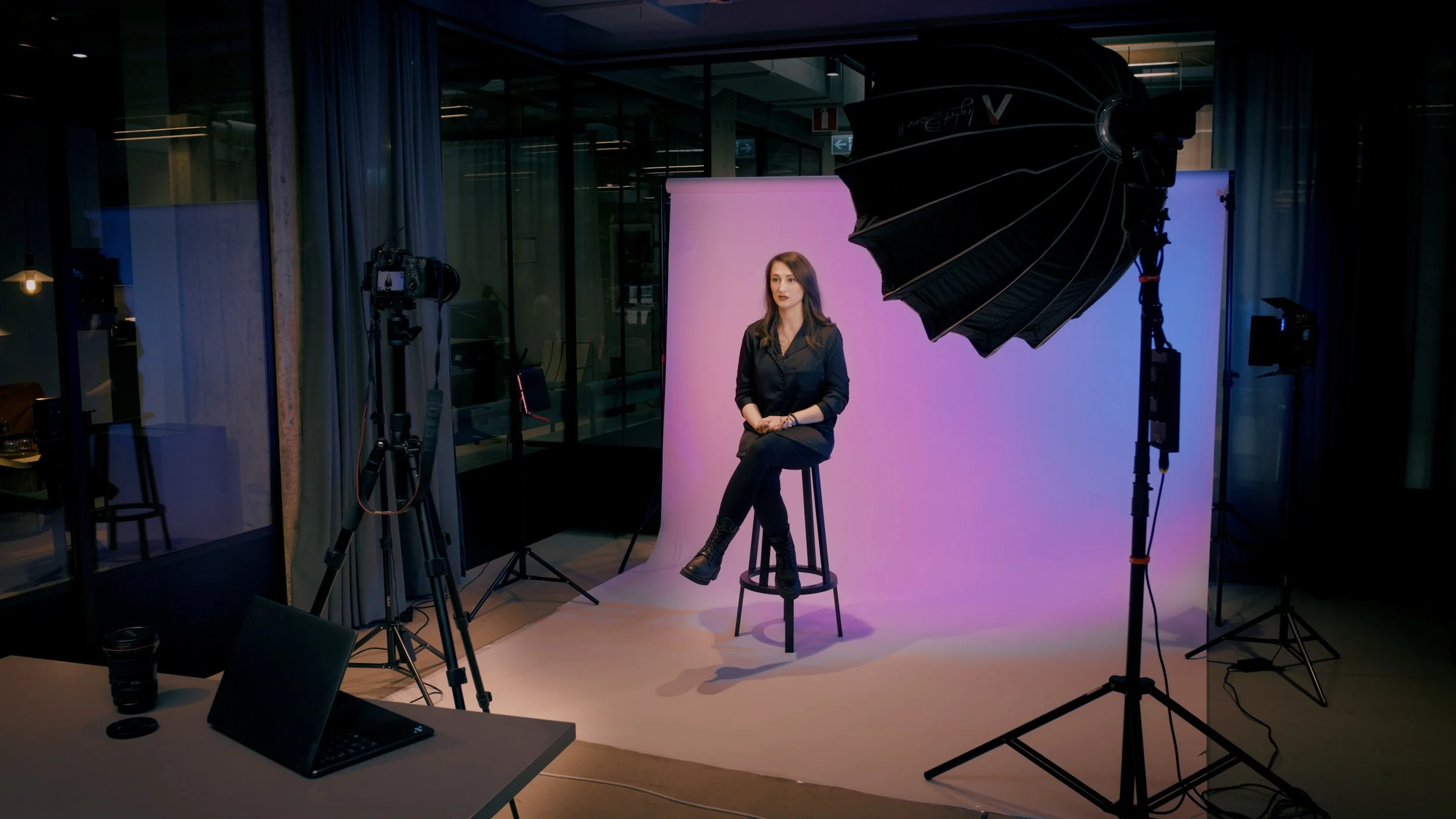 En kvinna sitter på en pall framför en rosa bakgrund i en inspelningsstudio, omgiven av fotografi- och videoutrustning, inklusive kamera, ljus och reflektorer, i ett modernt kontorslandskap.