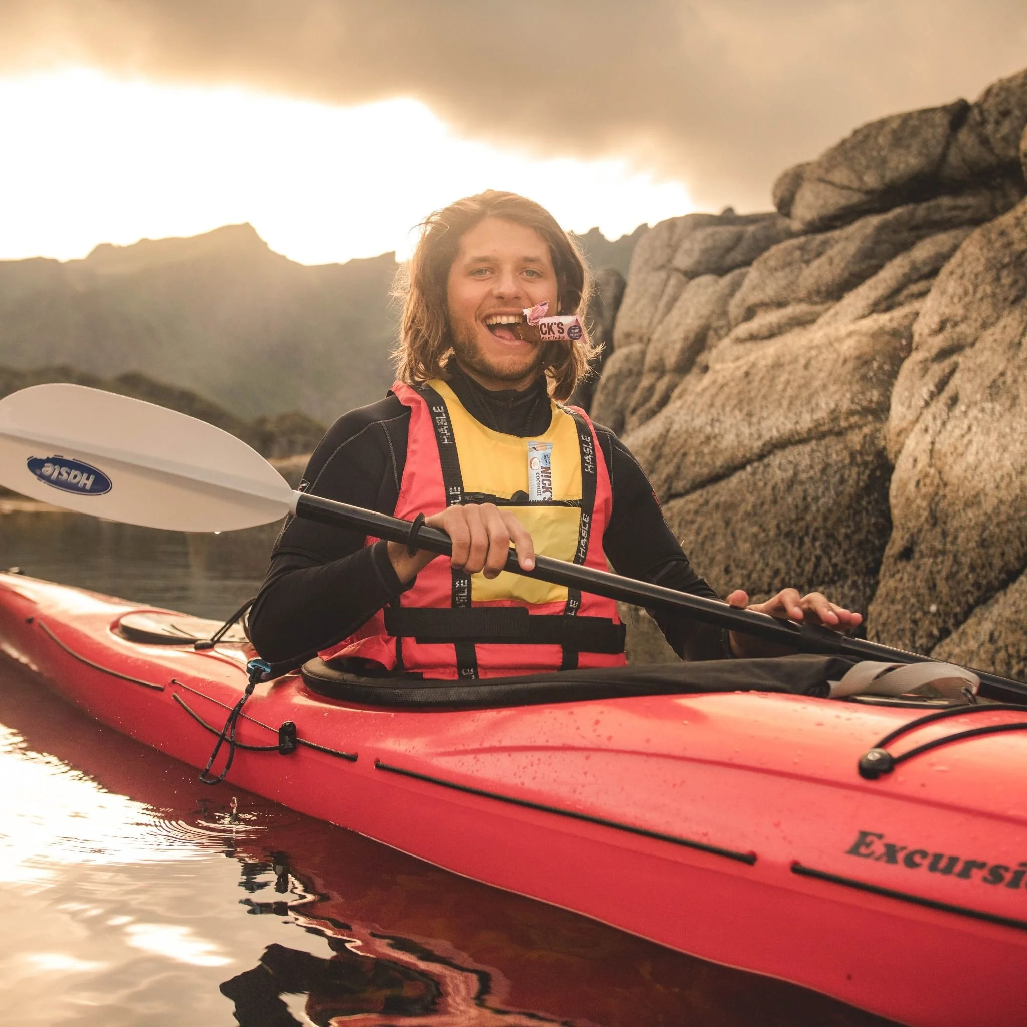 En man i en röd kajak på vattnet, med stenar och moln i bakgrunden. Han håller en paddel och verkar ha haft ett äventyr.