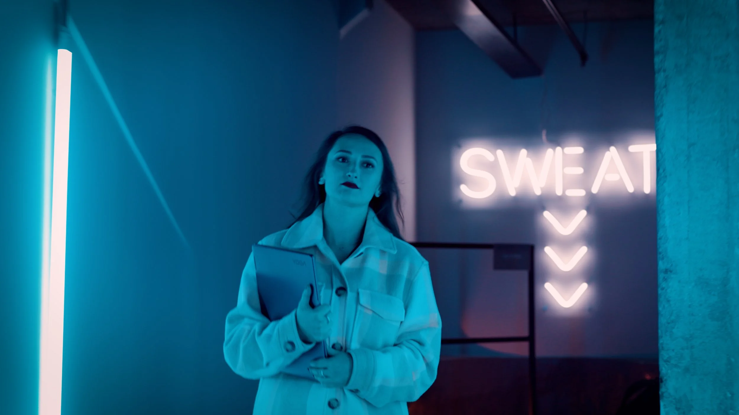 Kvinna står i ett rum med neonljus som säger 'SWEAT' och pilar nedåt, hon håller en surfplatta.