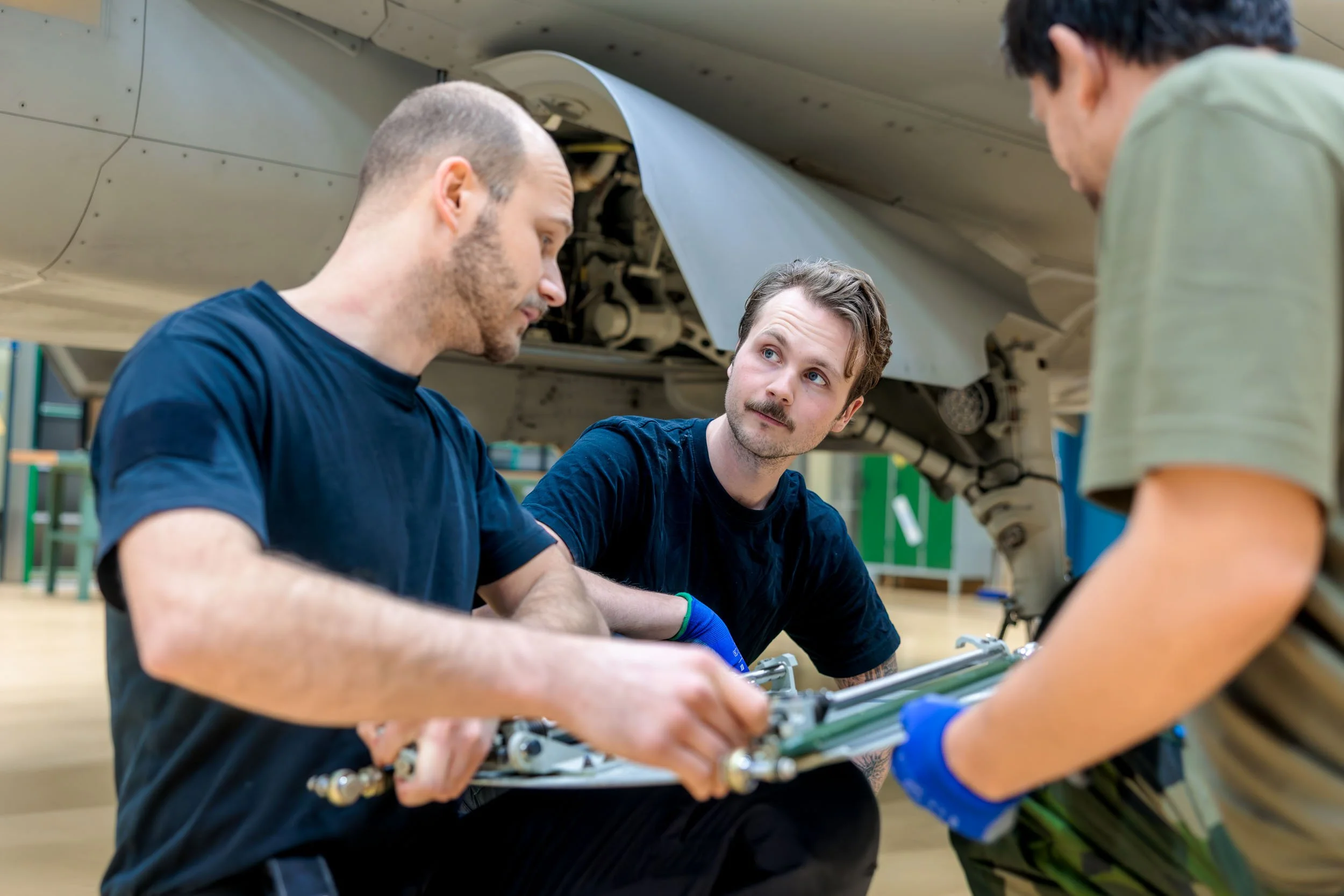 Tre personer arbetar på ett flygplan i ett hangar, chefen ger instruktioner medan de andra fixar med flygplansdelar.