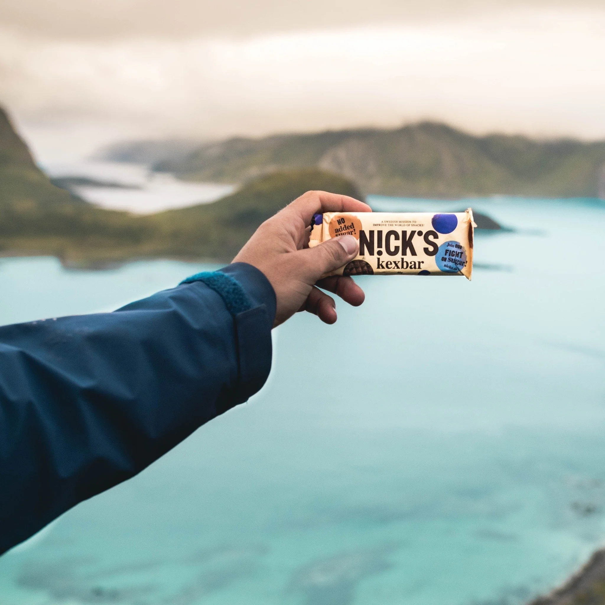 Person håller i en NICK'S kexbar framför en vattenlandskap med fjäll och sjöar.