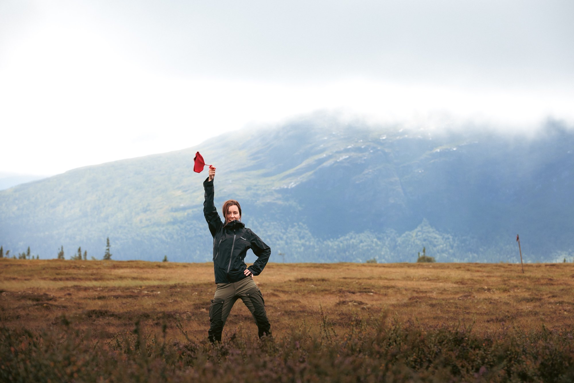 En kvinna står på en öppen äng, håller en röd flagga i luften, med berg och moln i bakgrunden.