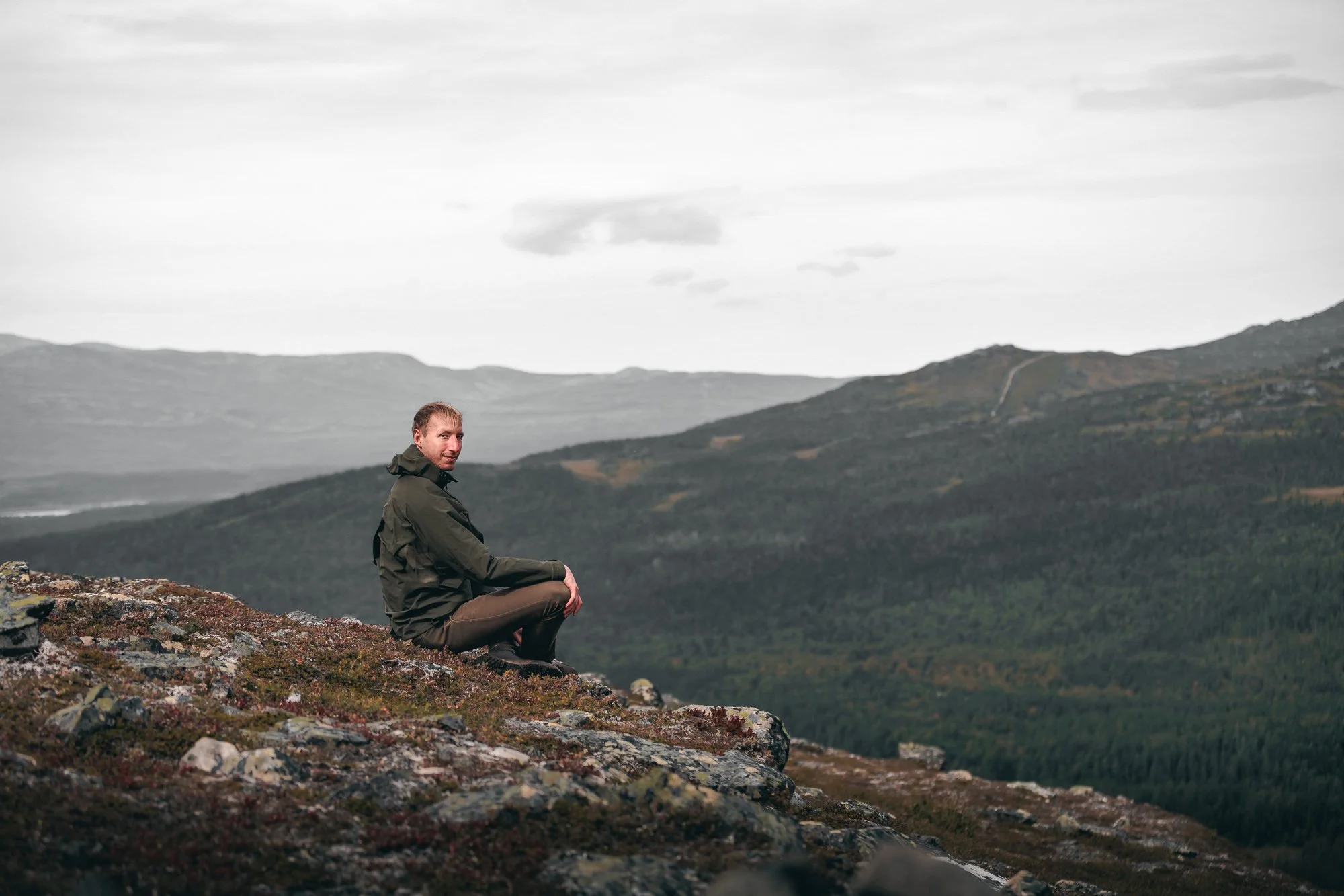 Man i gröna kläder som sitter på en sten i ett bergslandskap med gröna åsar och molnigt himmel.