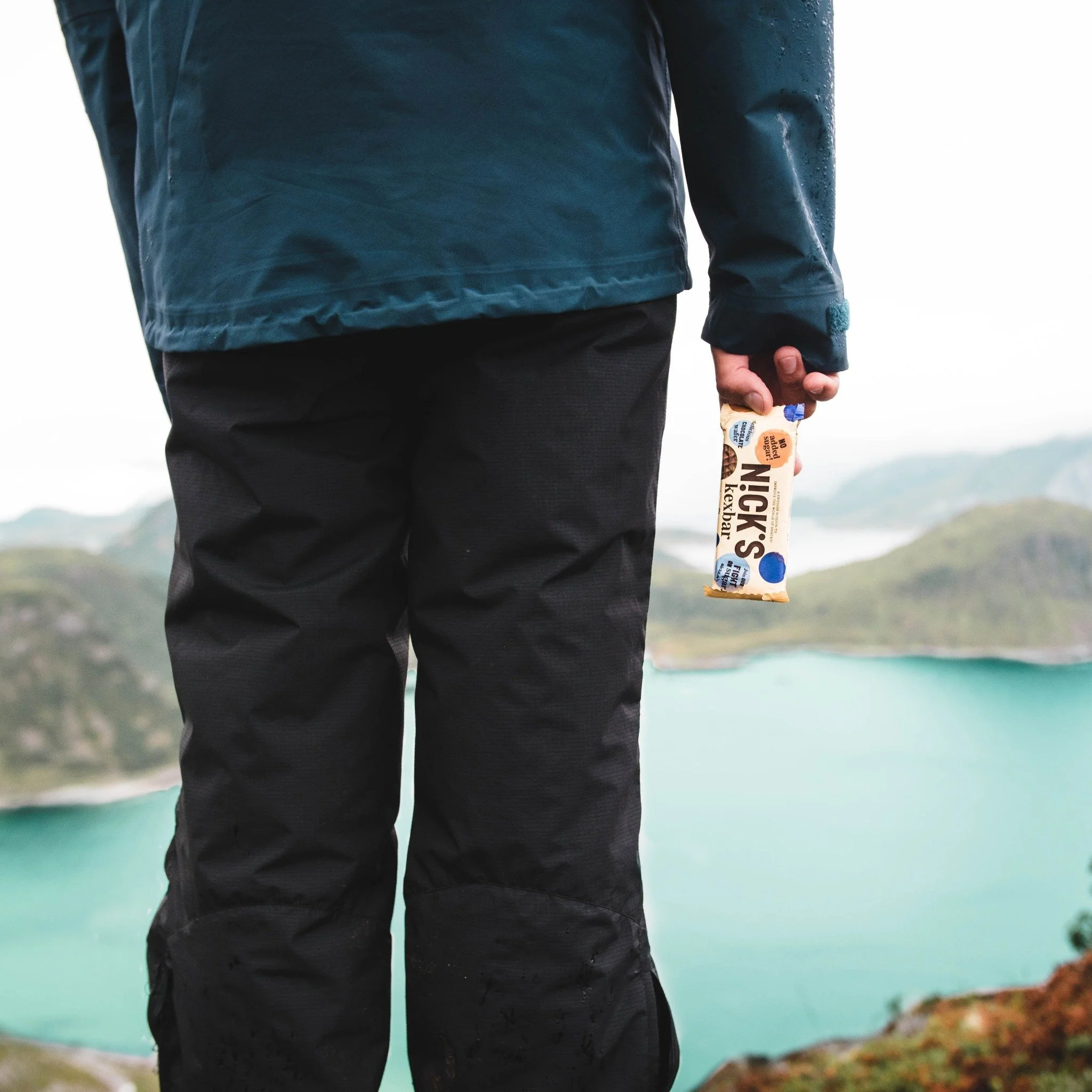 En person i vandrarkläder som håller i en Näcks traditionella knäckebrödspaket, med fjäll och sjö i bakgrunden.