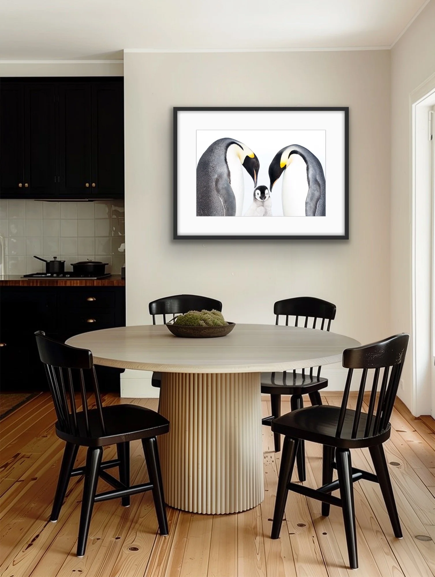 A framed photo of two adult penguins and a penguin chick hangs in a light dining room. The walls are cream, with a light, modern circular dining table and 4 black chairs in front. A modern, dark blue kitchen is shown in the background.