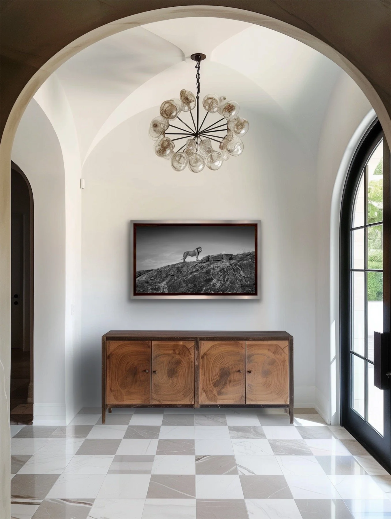 A black and white photo of a lion stood on a hill hangs in an entryway. The floor is pattern tiled, the walls are arched and a dark wood cabinet sits just below the framed photograph.