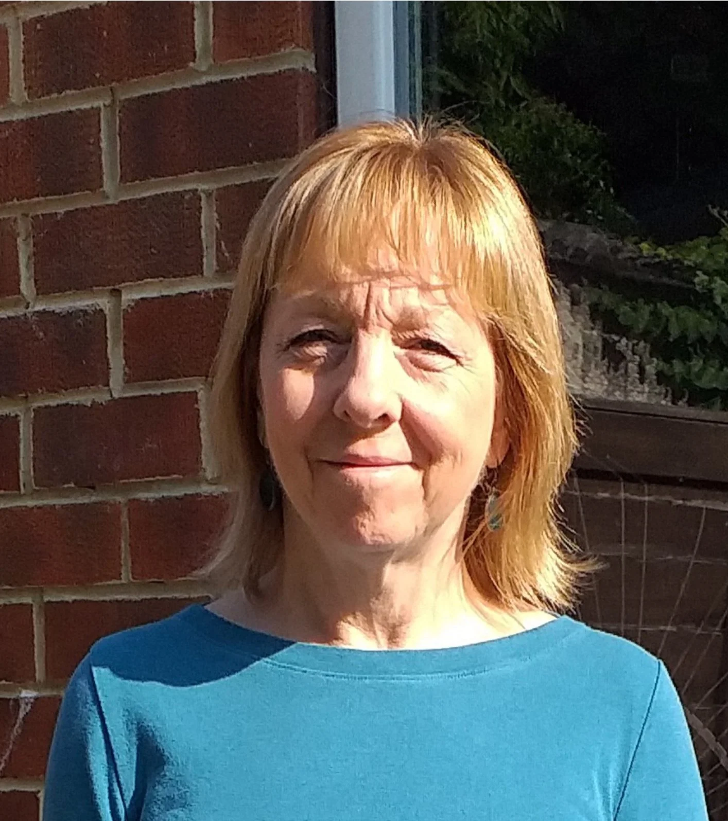 A woman with reddish-blonde hair standing outside in sunlight, smiling, near a brick wall and a bicycle.
