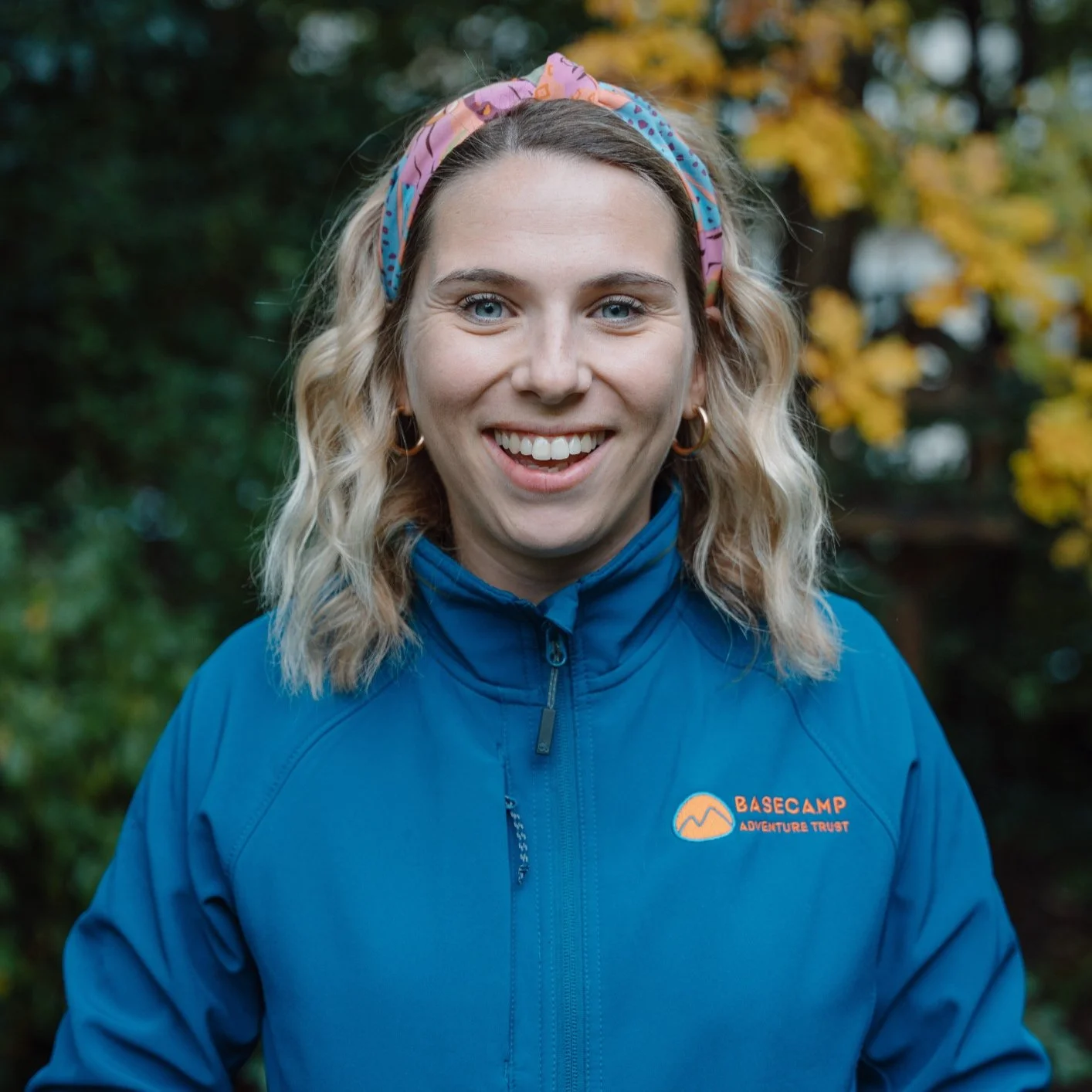 A smiling woman with blonde wavy hair wearing a pink and blue headband and a blue outdoor jacket in front of autumn trees with yellow leaves.