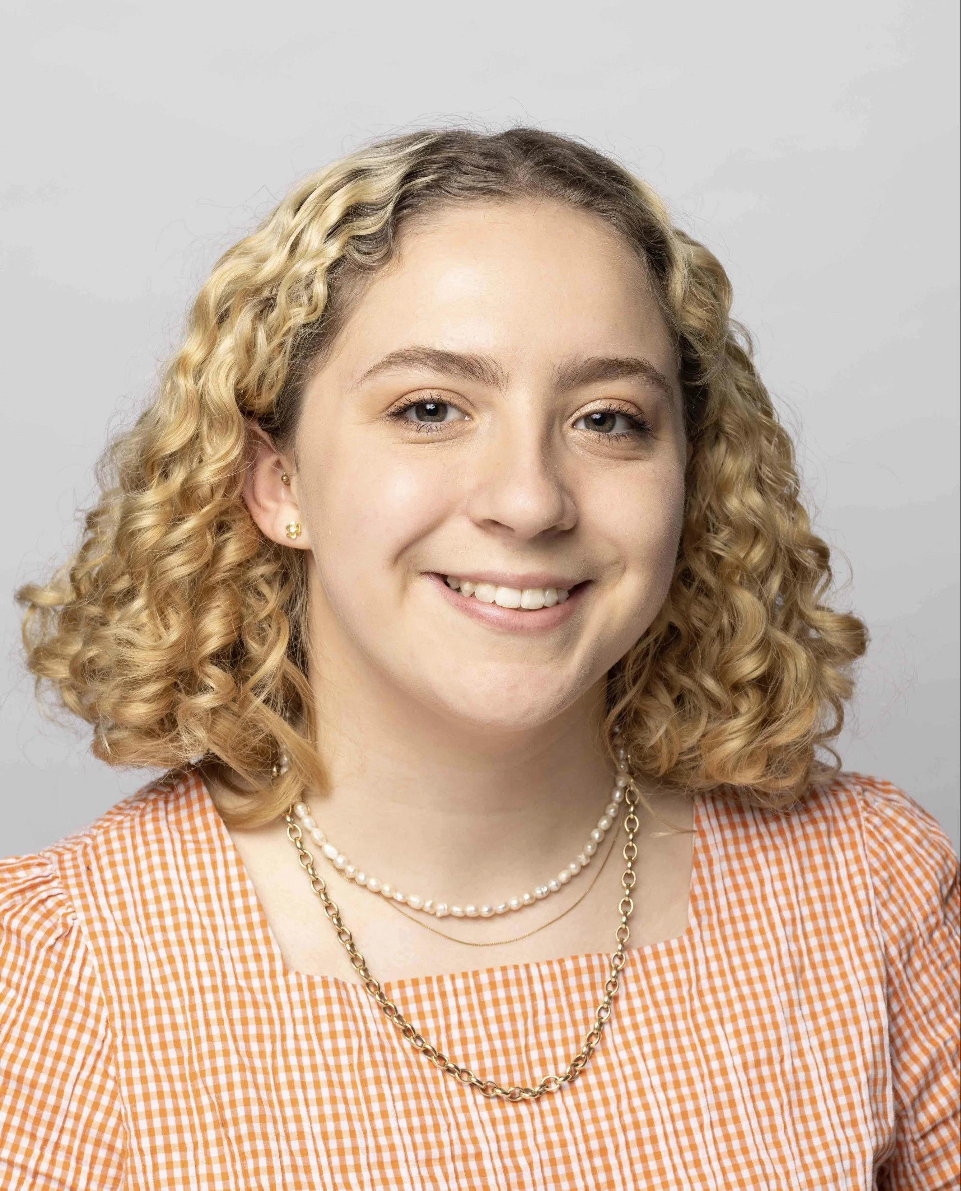 A woman with blonde, curly hair, wearing a pearl necklace and gold earrings, smiling, in an orange and white gingham top, against a light grey background.