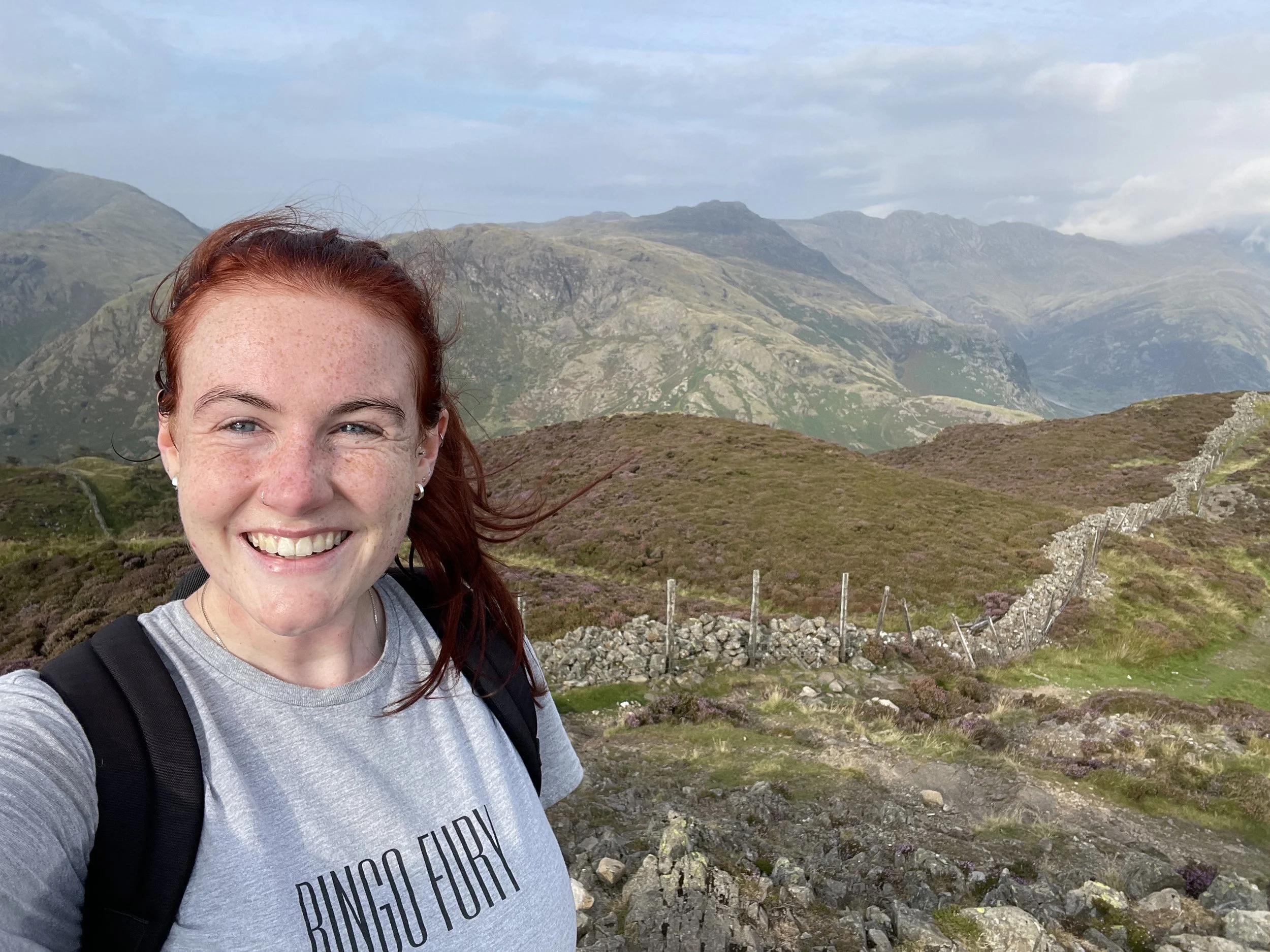 Smiling woman with red hair and freckles taking a selfie in a mountainous landscape with green hills, rocks, and cloudy sky in the background.