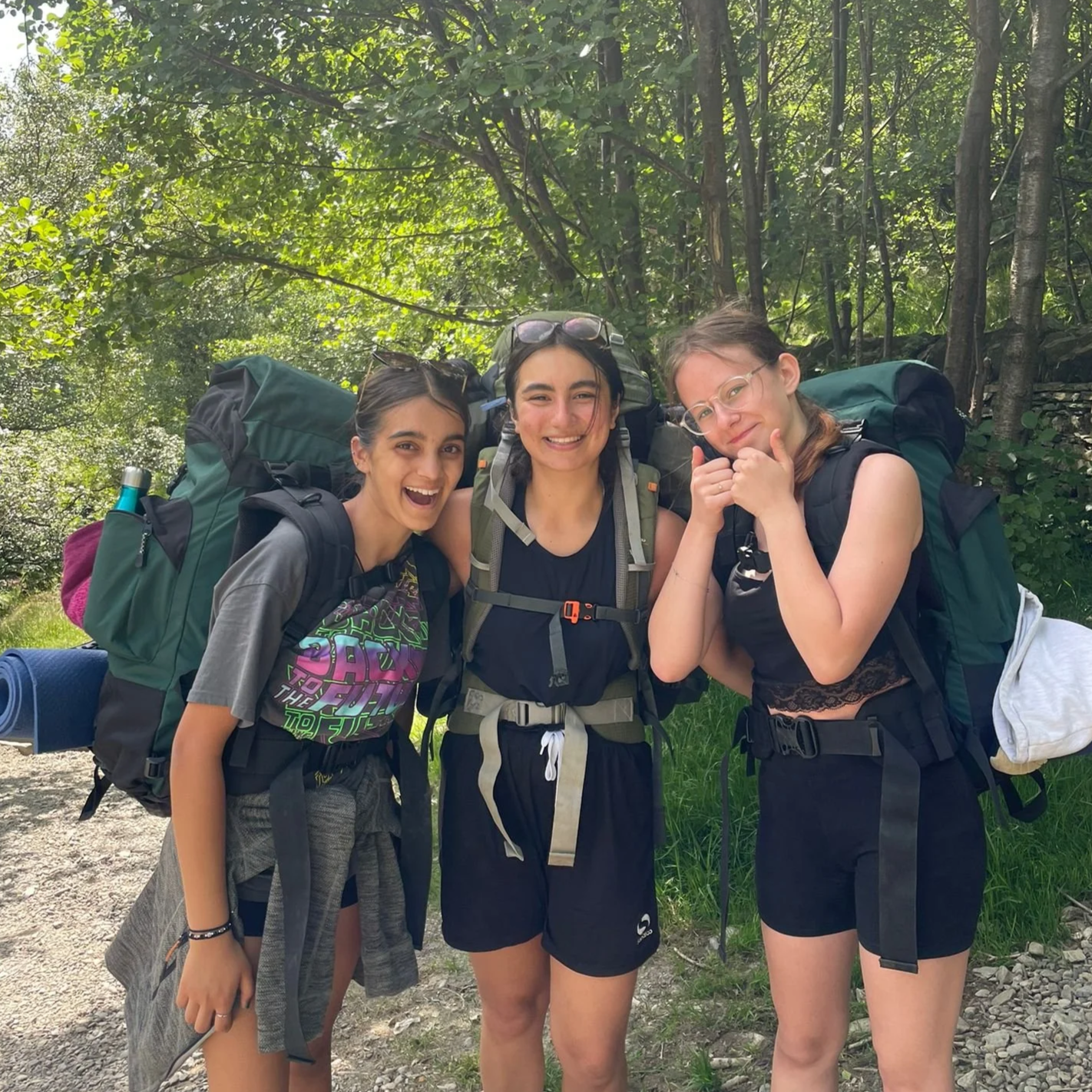 Three young women with backpacks smiling and posing for a photo outdoors in a forested area under green trees.