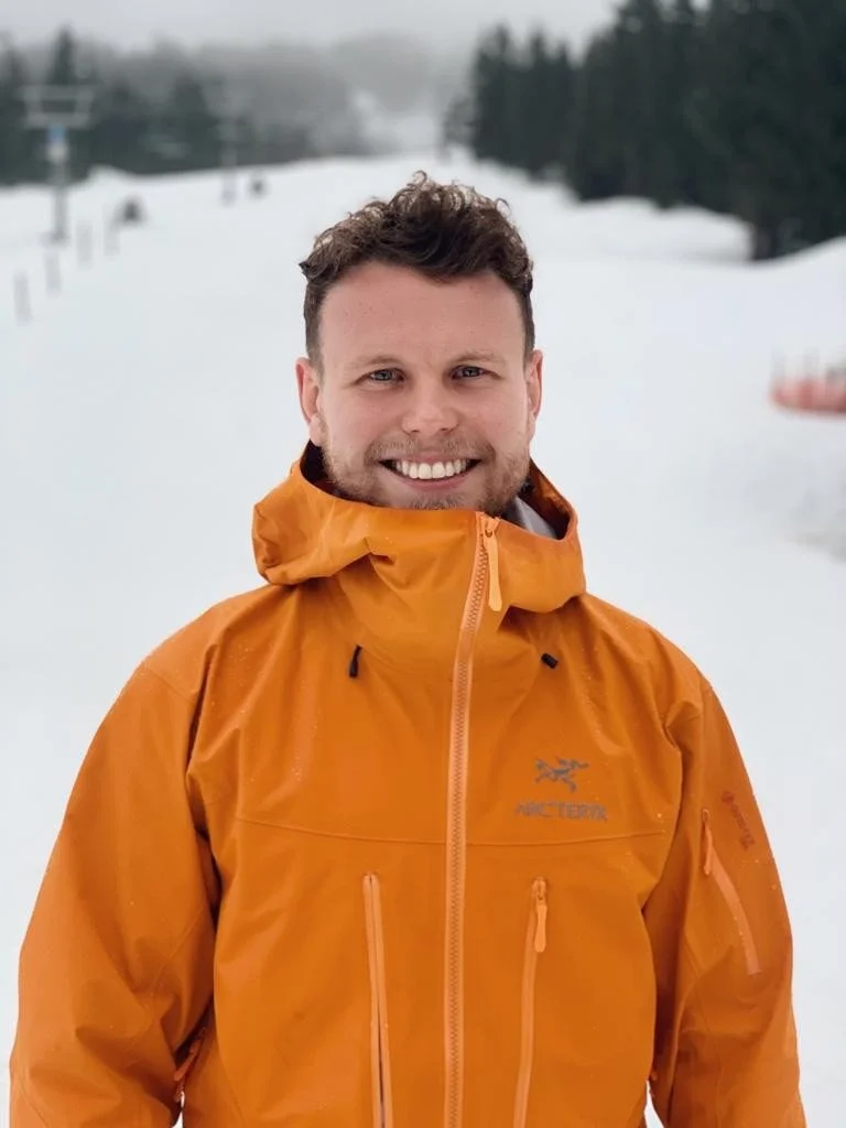 Smiling man in an orange Arc'teryx jacket standing outdoors on a snow-covered slope with trees and ski lift in the background.