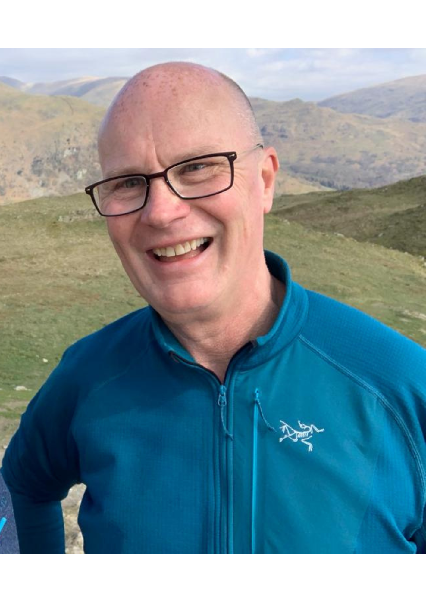 A man with glasses and a bald head smiling outdoors with rolling green hills and mountains in the background, wearing a blue jacket with a white dinosaur logo.