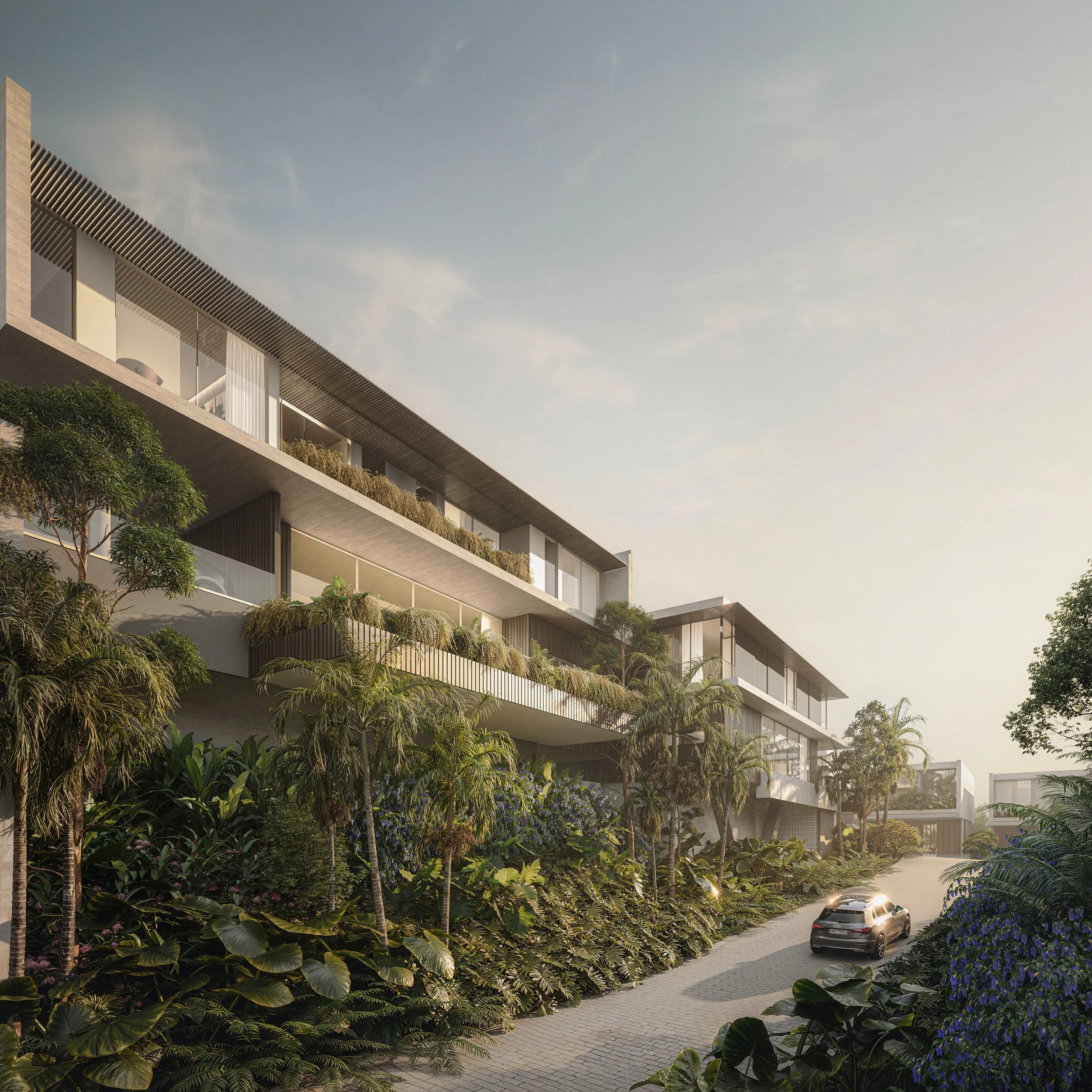 Modern multi-story apartment building with large glass windows and balconies, surrounded by lush green tropical plants and trees, with a paved driveway and a black car parked outside, under a clear sky with the sun setting.