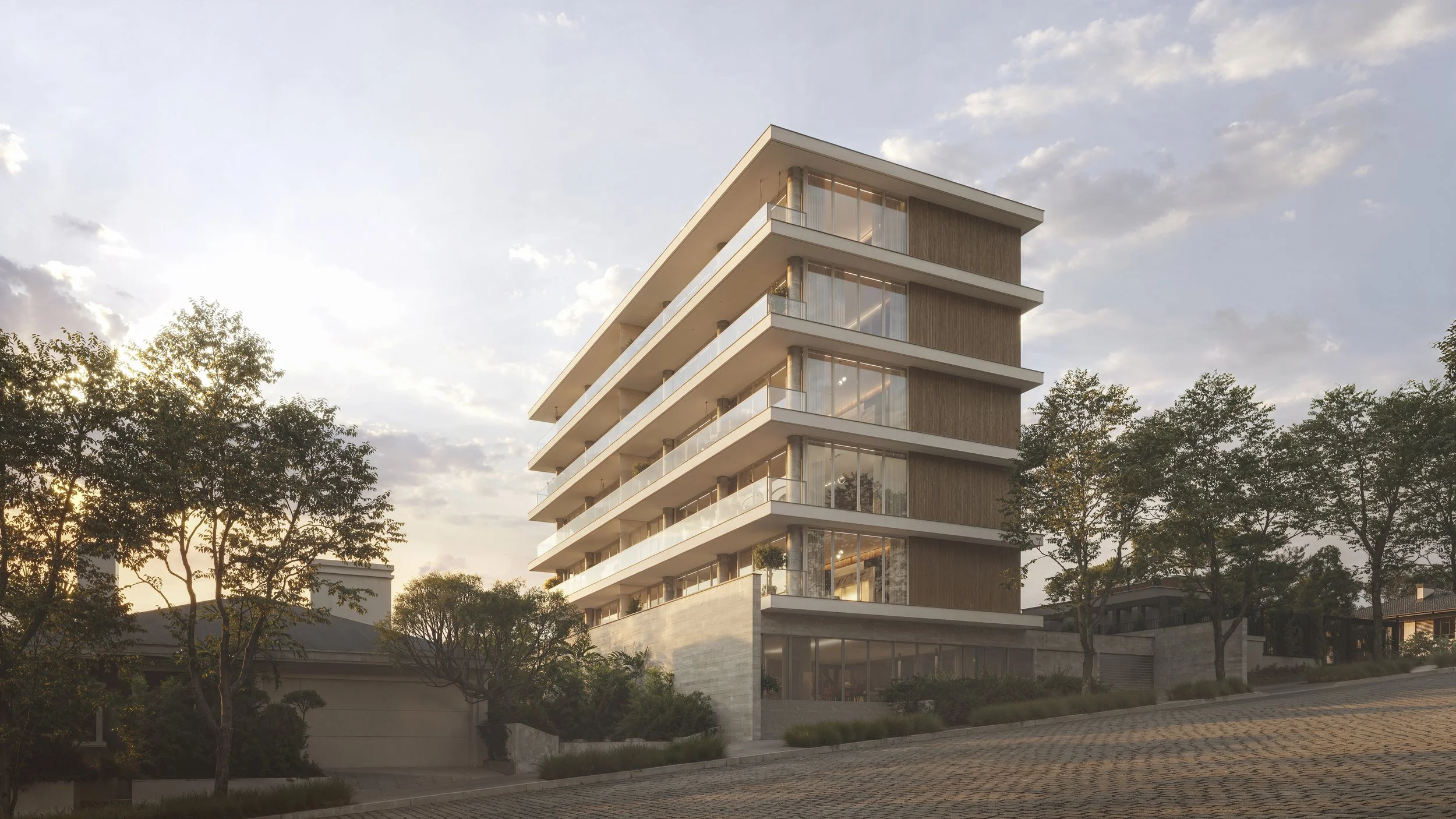 A modern multi-story residential building with large glass windows, situated on a slightly sloped street with trees and greenery surrounding it, during sunset.