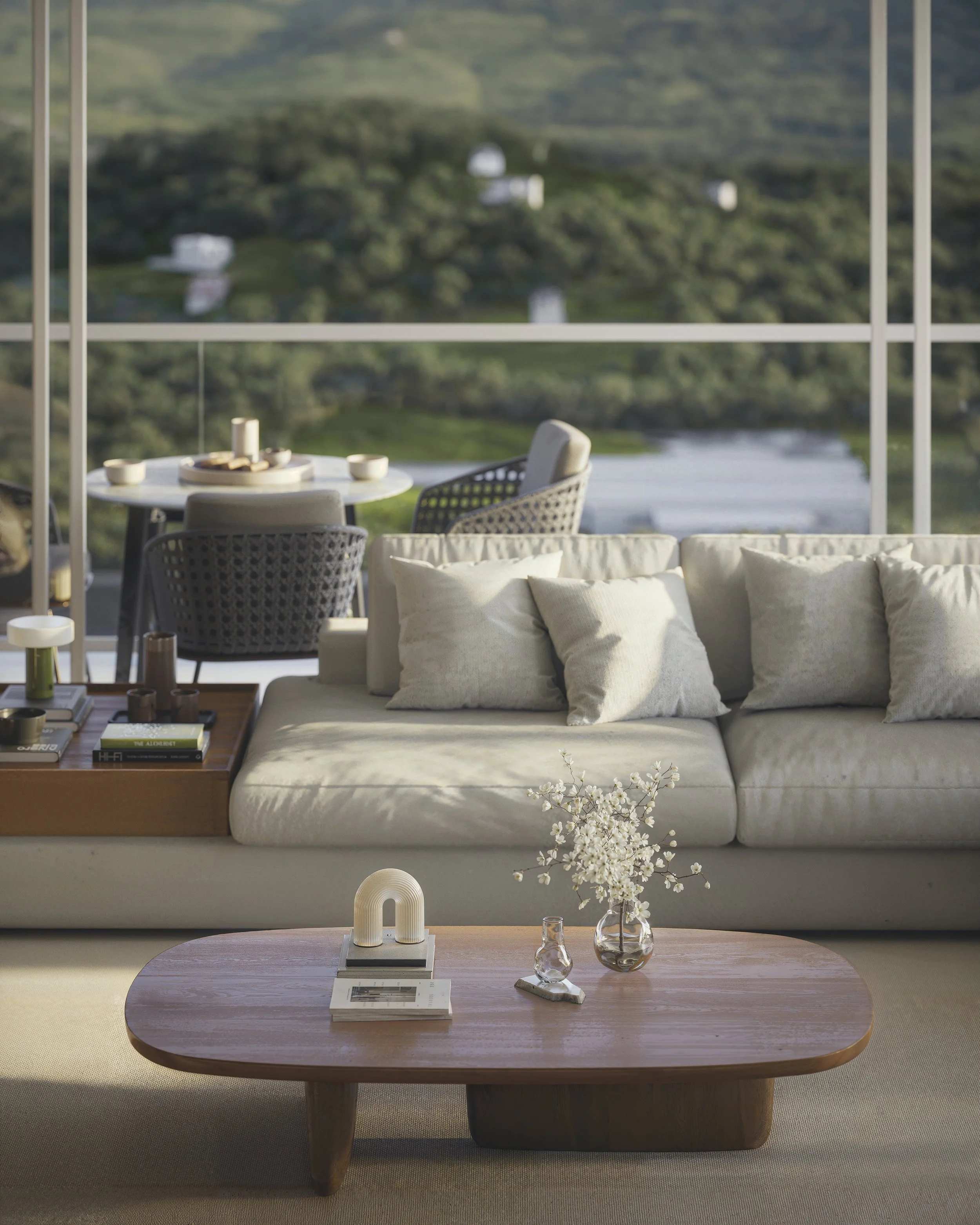Living room with a white sofa and wooden coffee table, decorated with vases and flowers, overlooking an outdoor patio with tables and chairs, and a scenic green landscape in the background.