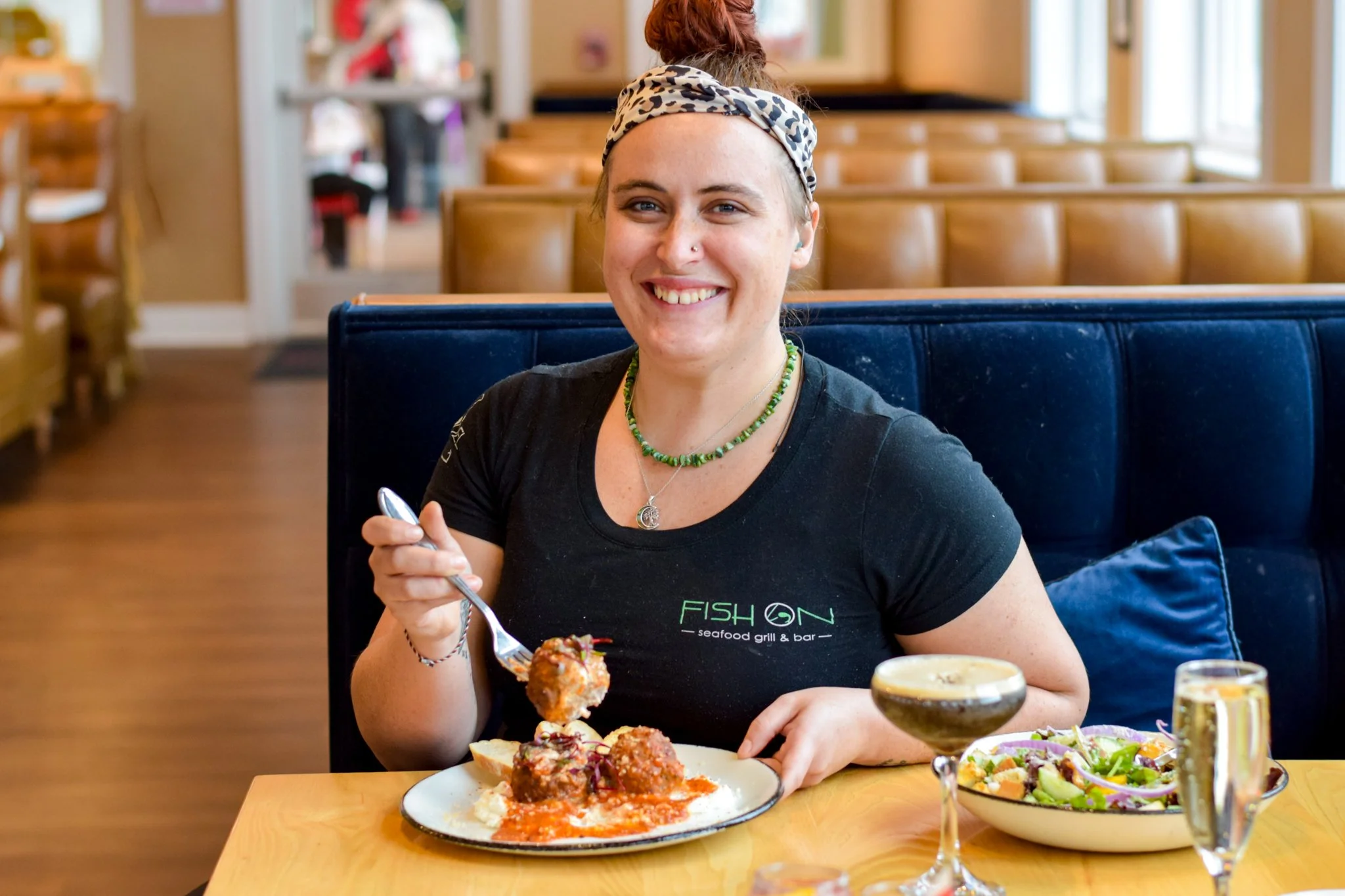 Chef Maggie Williamson, smiling and holding a fork, sitting at a table with a plate of meatballs, a bowl of salad, and drinks in a restaurant with wooden furniture.
